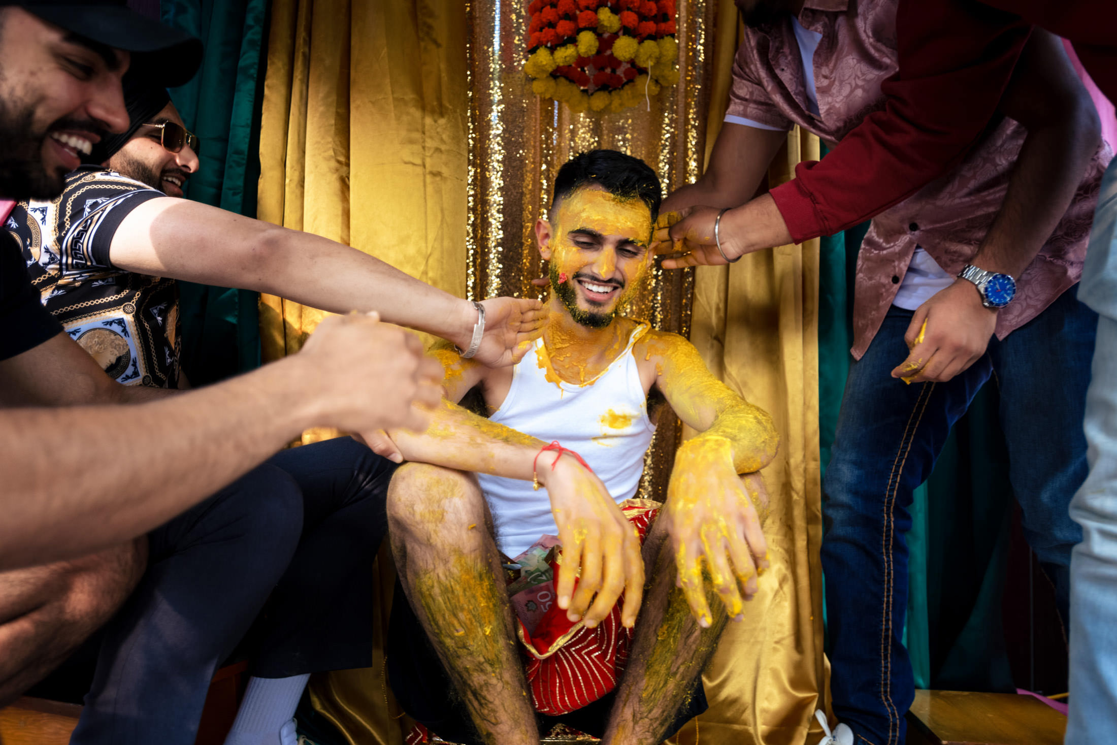 Man sitting, covered in yellow paste, smiling as friends apply more during a Winnipeg wedding.