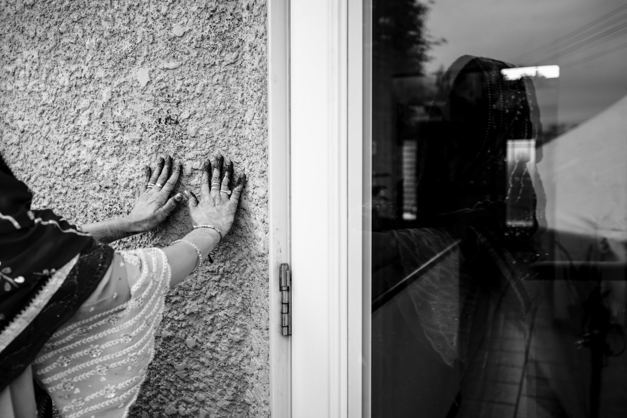 Bride in traditional attire presses hands on wall, reflection in glass, Winnipeg wedding vibes.