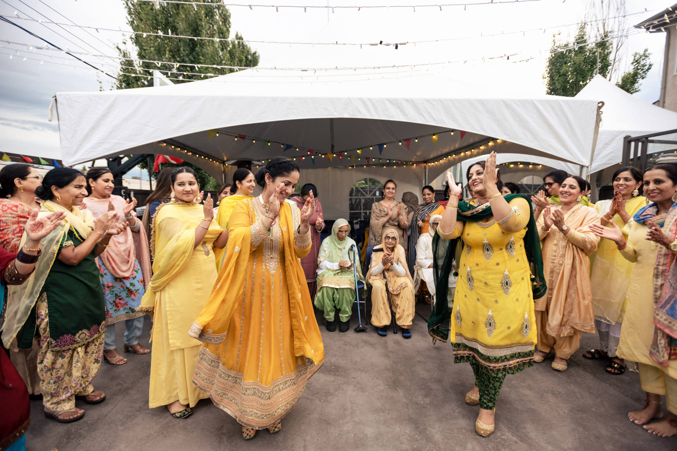 People in colorful traditional clothing dance joyfully at a Winnipeg wedding under string lights.