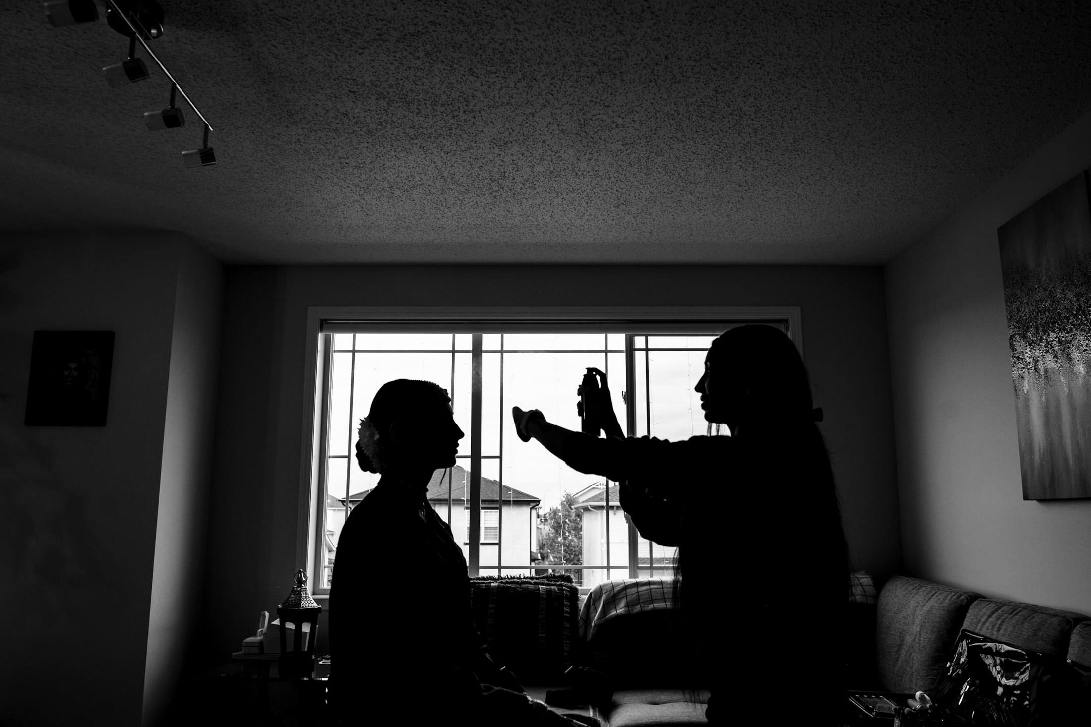 Silhouettes at a window; one seated, another with hair spray, capture a Winnipeg wedding moment.