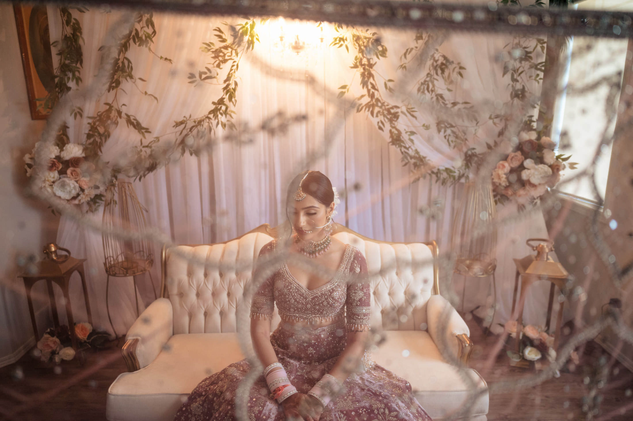 Bride in ornate attire, seated on sofa with floral backdrop, viewed through Winnipeg wedding glass.