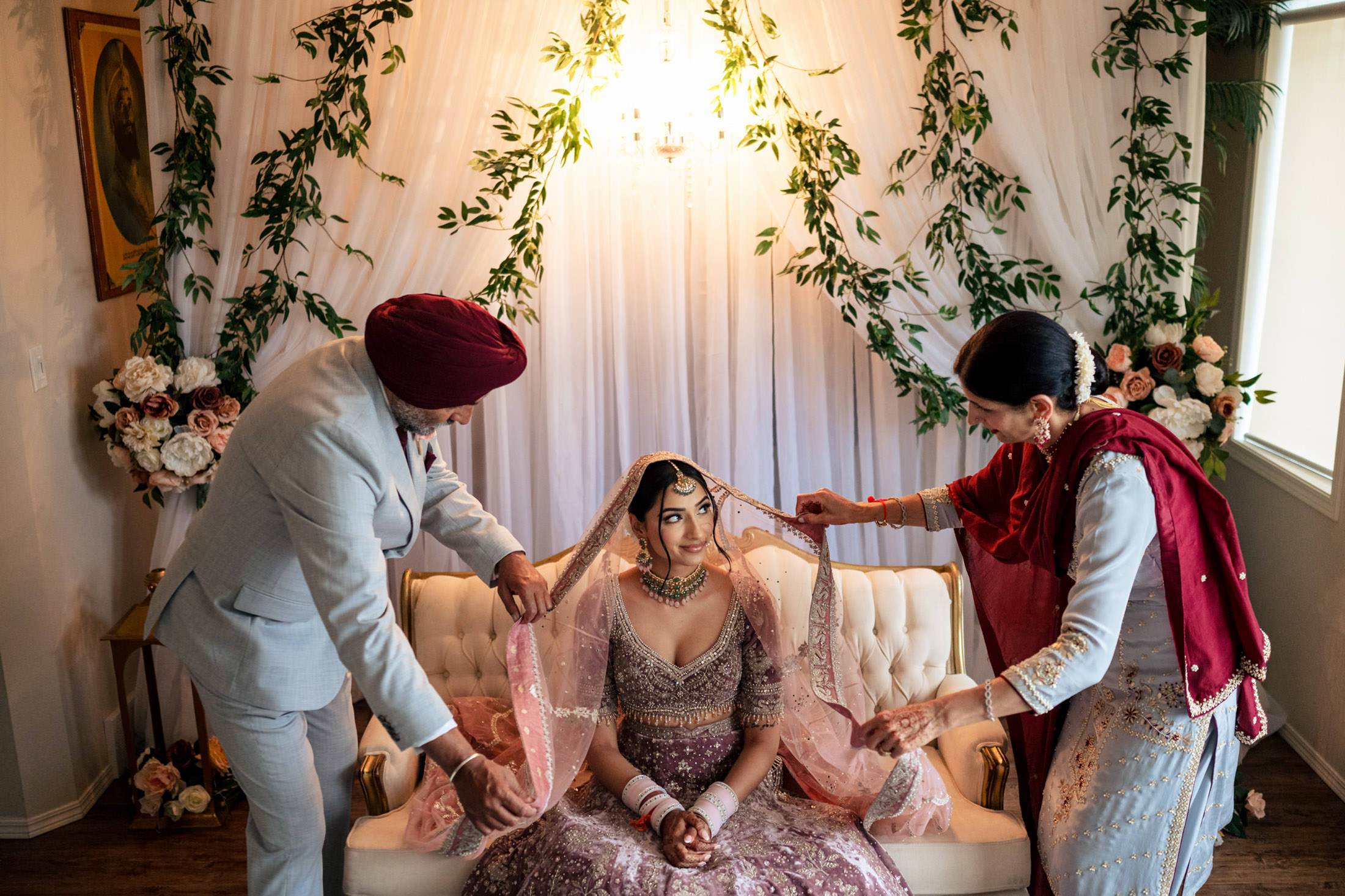 In a Winnipeg wedding, a bride sits as two people adjust her veil in a decorated room.