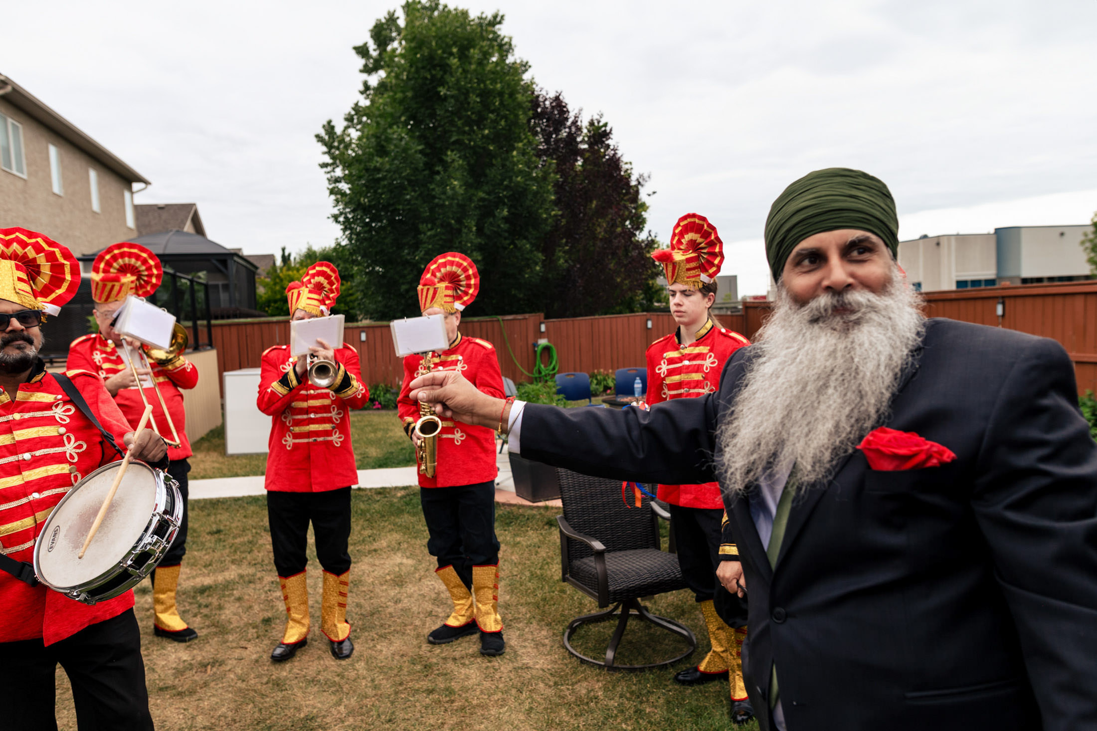 Man with beard gestures by a Winnipeg marching band in red uniforms outdoors.