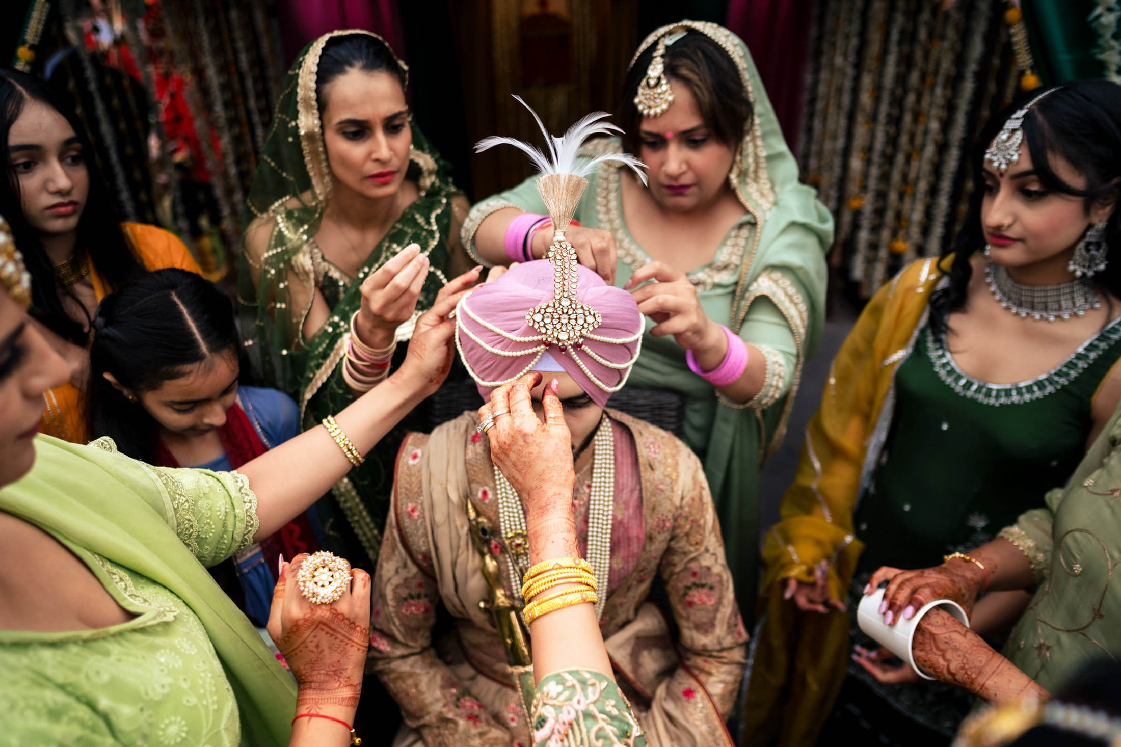 People in vibrant traditional attire adjust a groom's pink turban at a Winnipeg wedding.