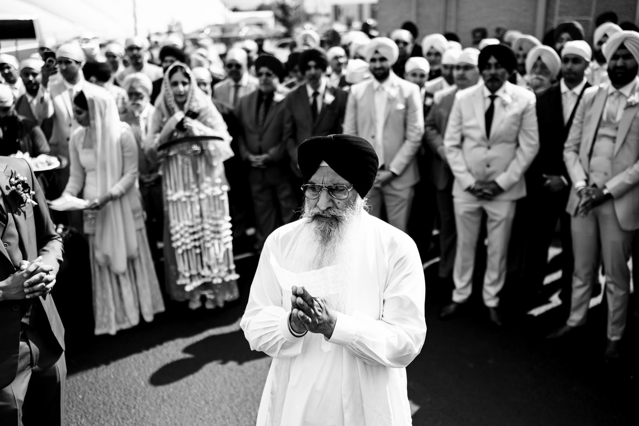 Elderly man praying at an outdoor Winnipeg wedding, surrounded by a crowd in traditional attire.