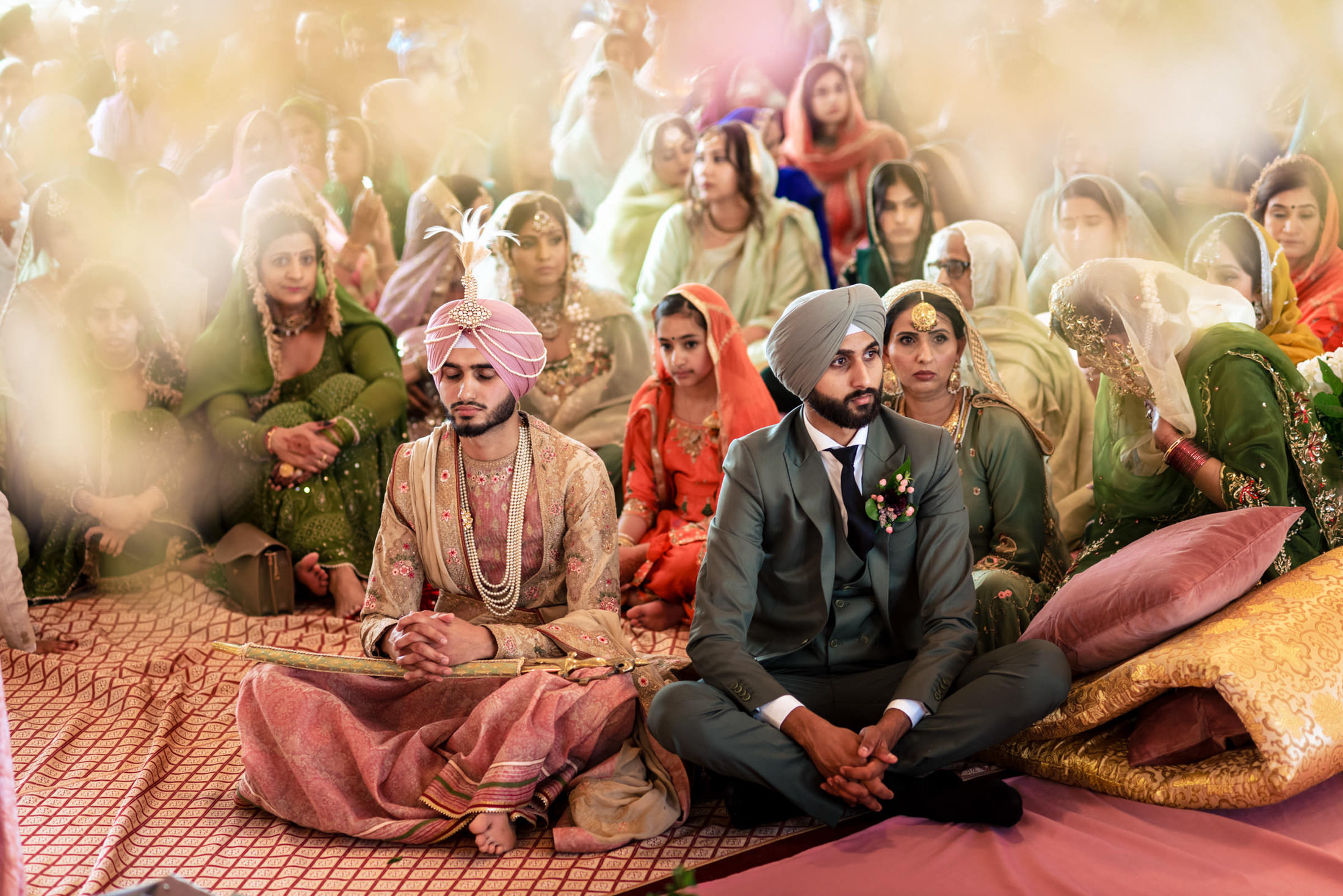 Two grooms sit cross-legged amid a colorful crowd at a vibrant Winnipeg wedding ceremony.