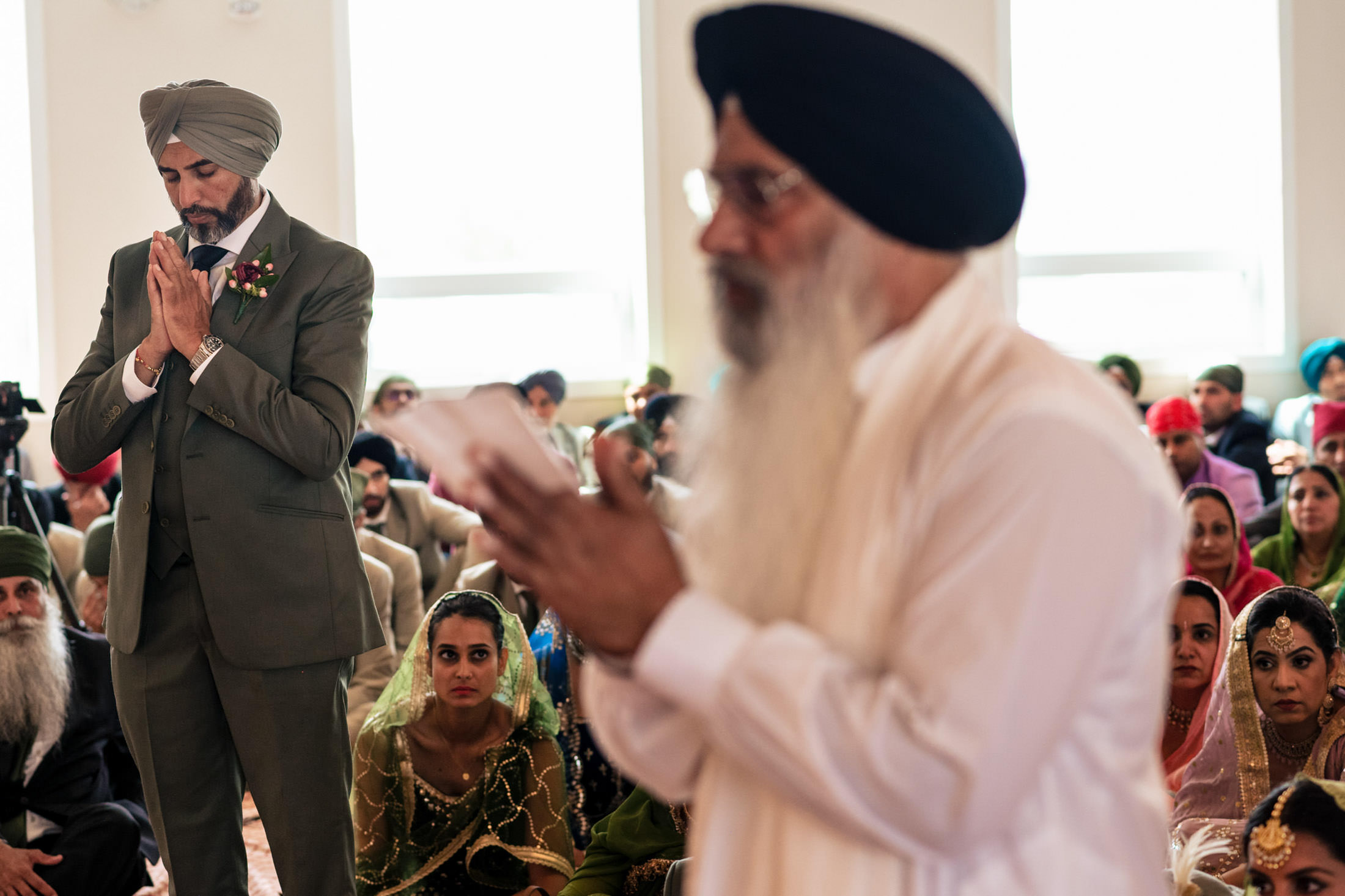Man in green suit praying at a Sikh ceremony, Winnipeg wedding shown in the background.