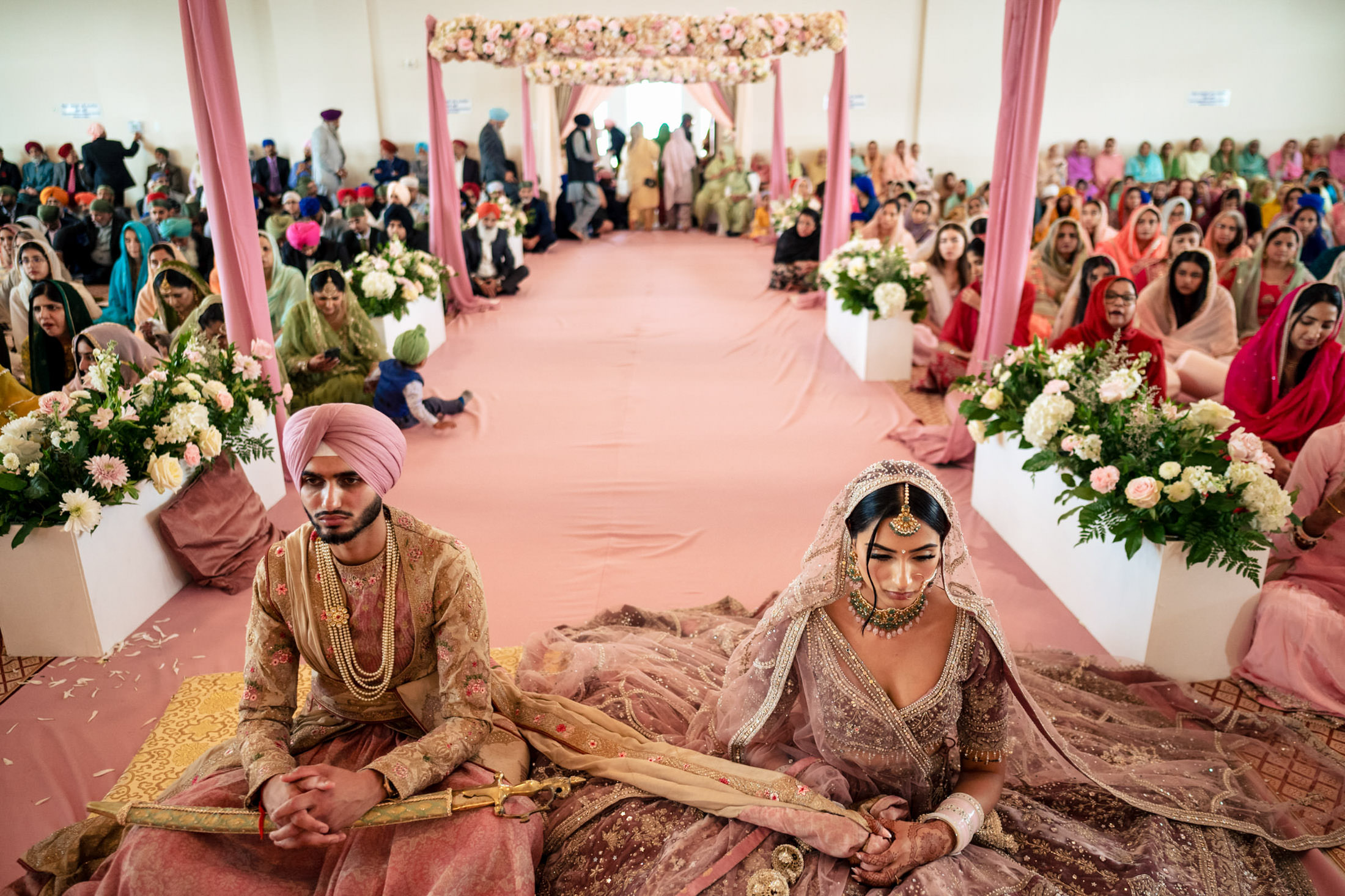 A couple in Winnipeg sits in traditional attire at a vibrant, floral-decorated wedding ceremony.