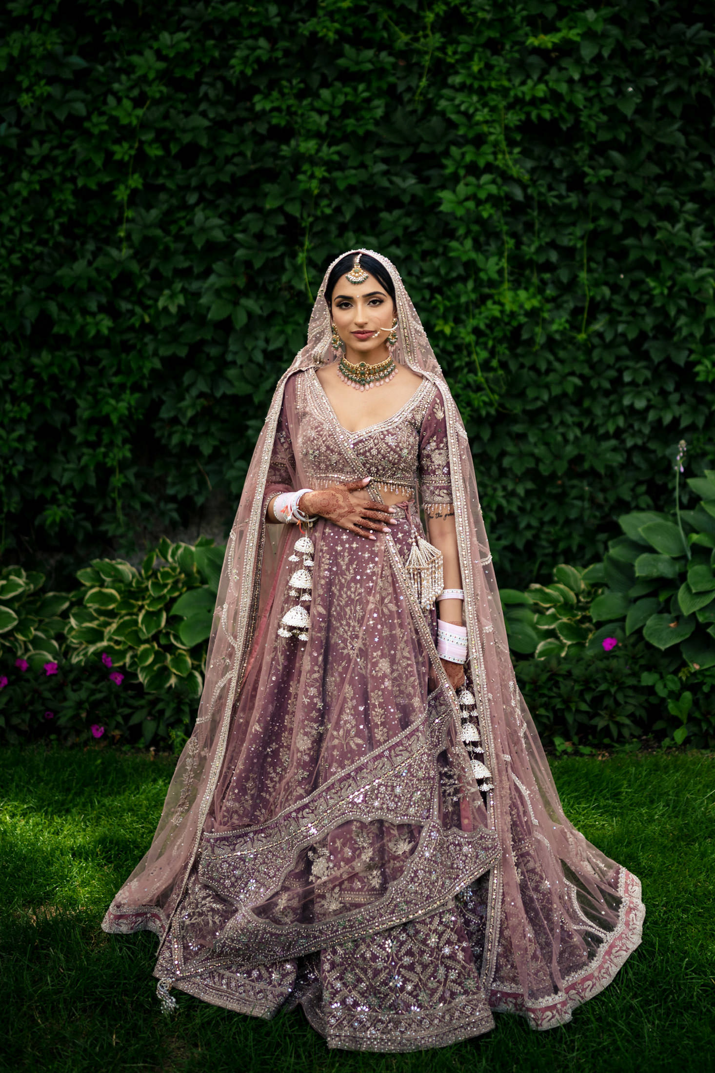 Bride in a detailed purple lehenga stands before a leafy backdrop at a Winnipeg wedding.