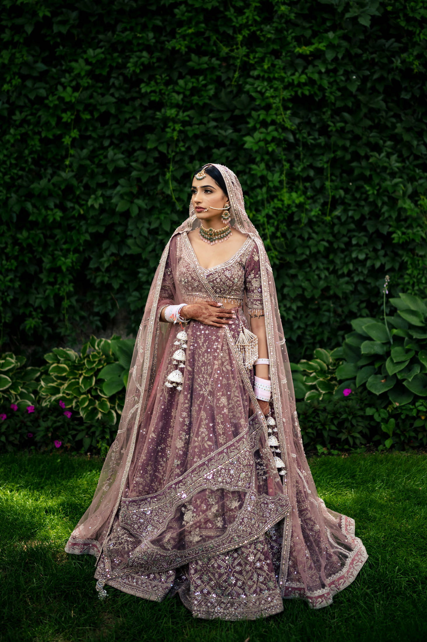 Bride in ornate pink lehenga poses against lush Winnipeg wedding greenery.