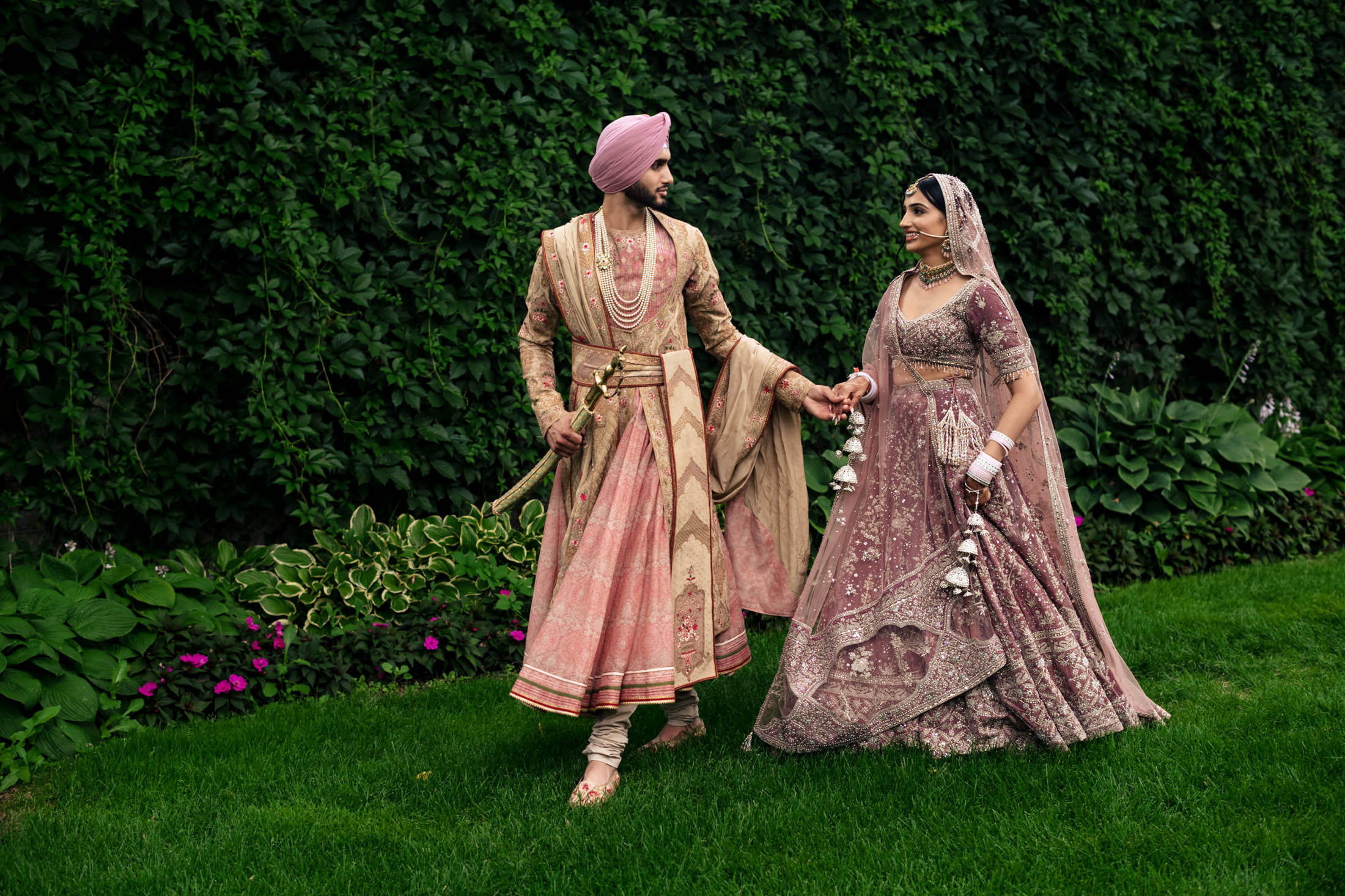 Couple in traditional attire stroll hand in hand at a Winnipeg wedding with a leafy backdrop.