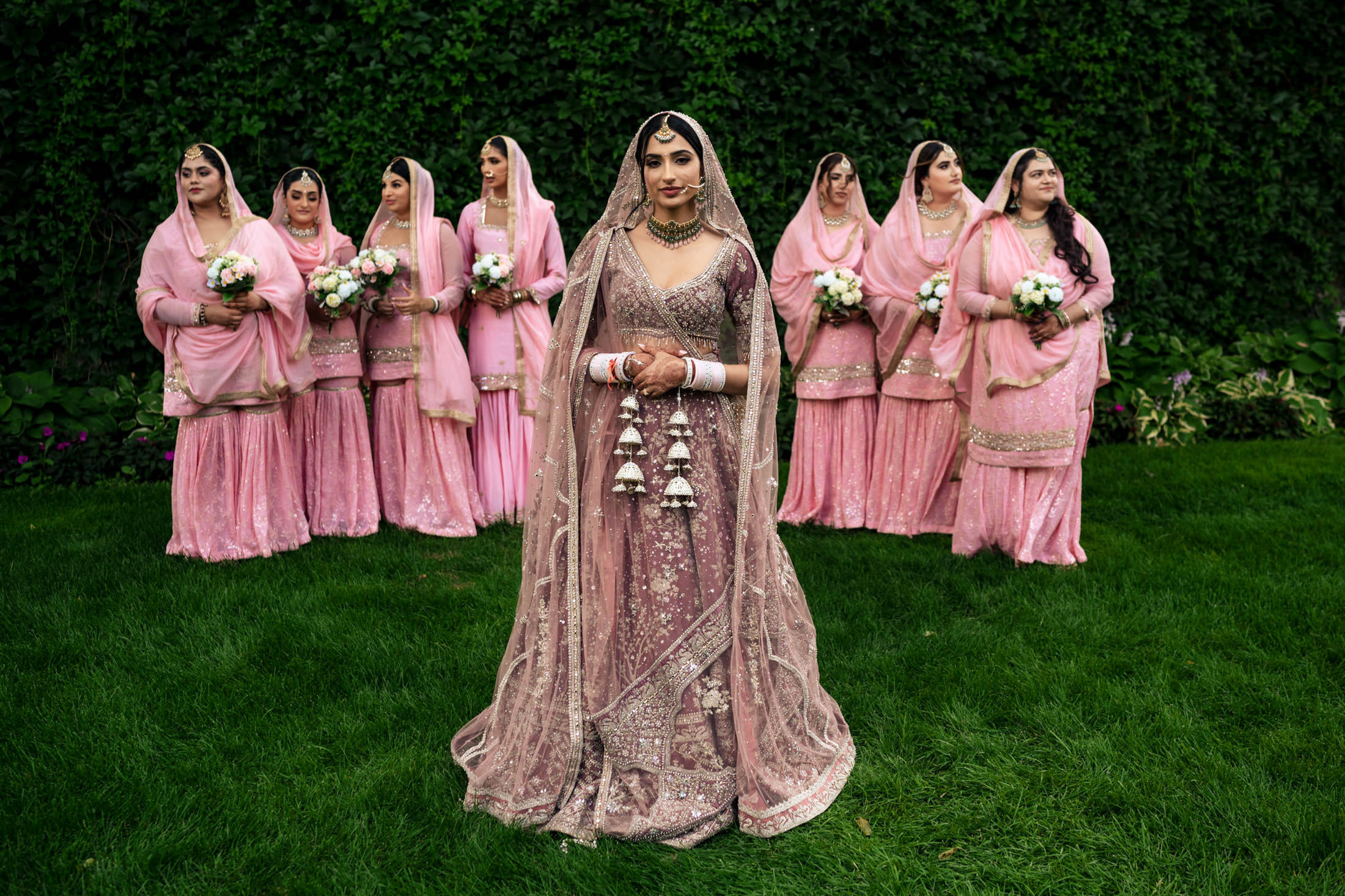 Bride in ornate dress stands before bridesmaids in pink at scenic Winnipeg wedding.