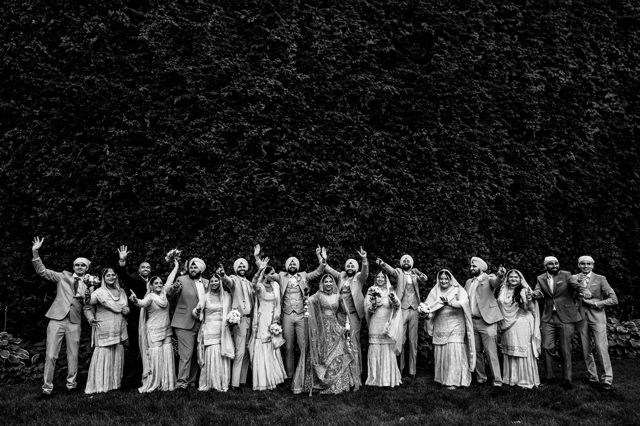 A large Winnipeg wedding party in traditional attire poses cheerfully amid a leafy backdrop.