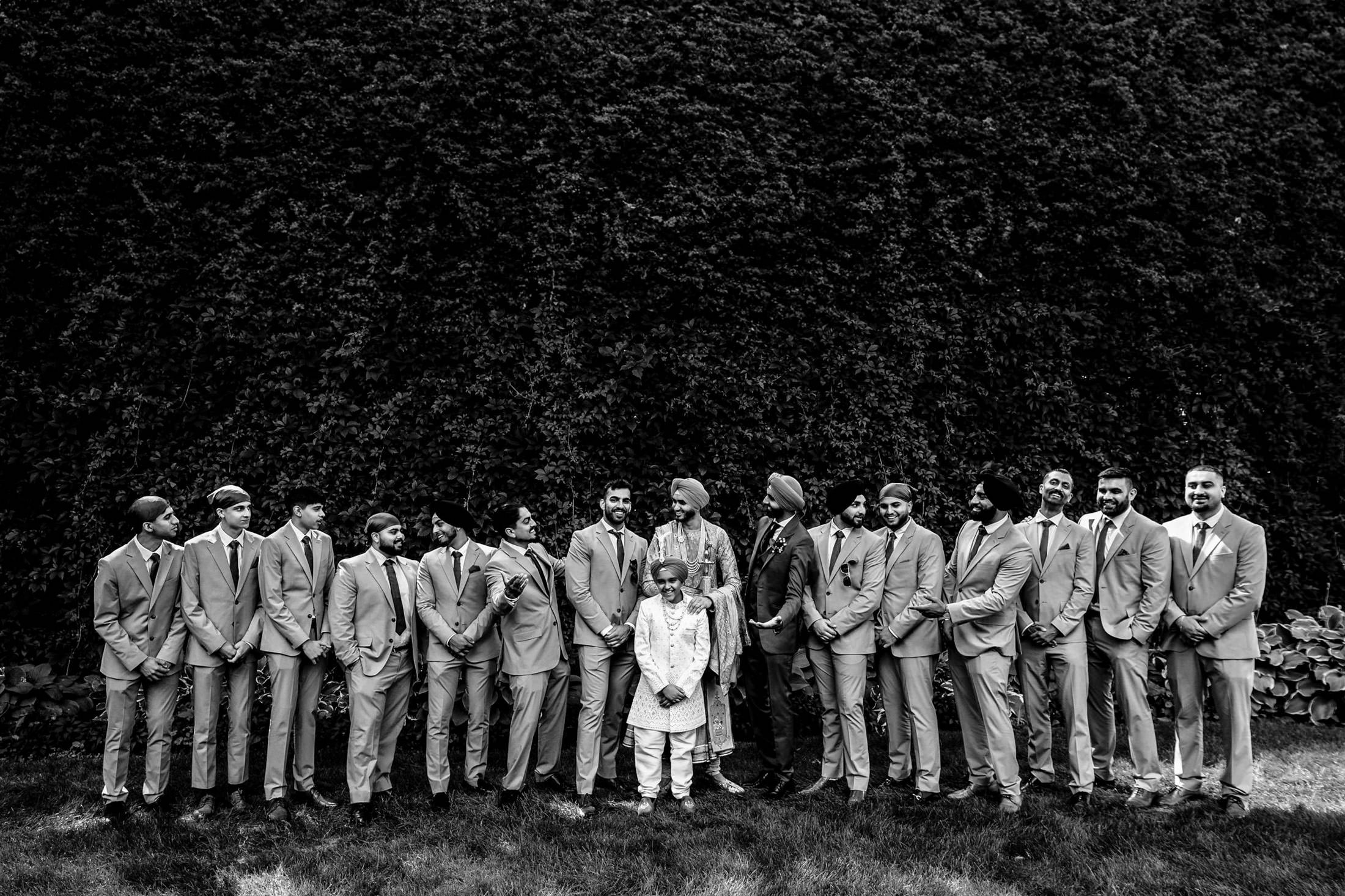 Group of men and a young child in suits, ready for a Winnipeg wedding by a leafy hedge.