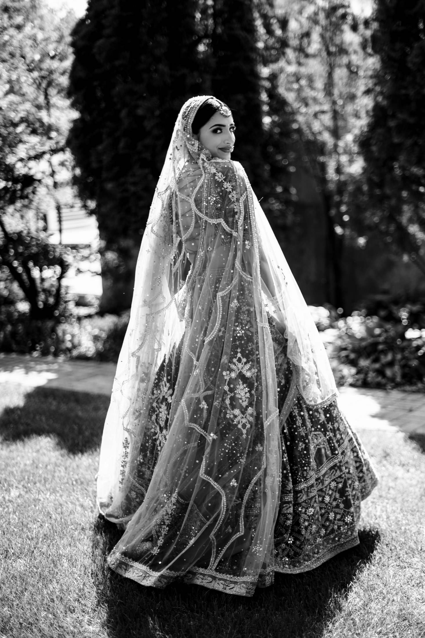 Bride in ornate dress and veil on grass, looking back over her shoulder at Winnipeg wedding.