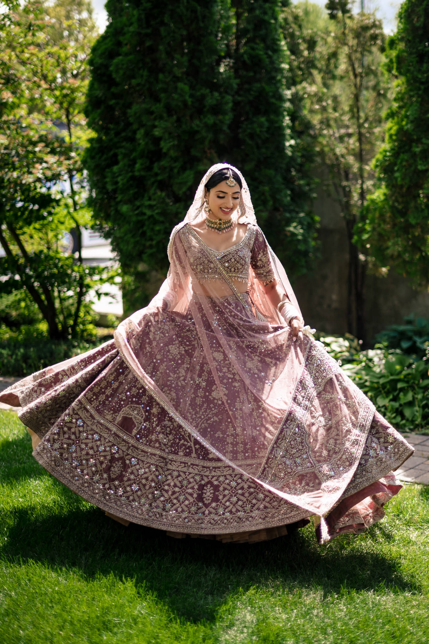 Woman in ornate lehenga twirling on grass at a Winnipeg wedding with trees behind.
