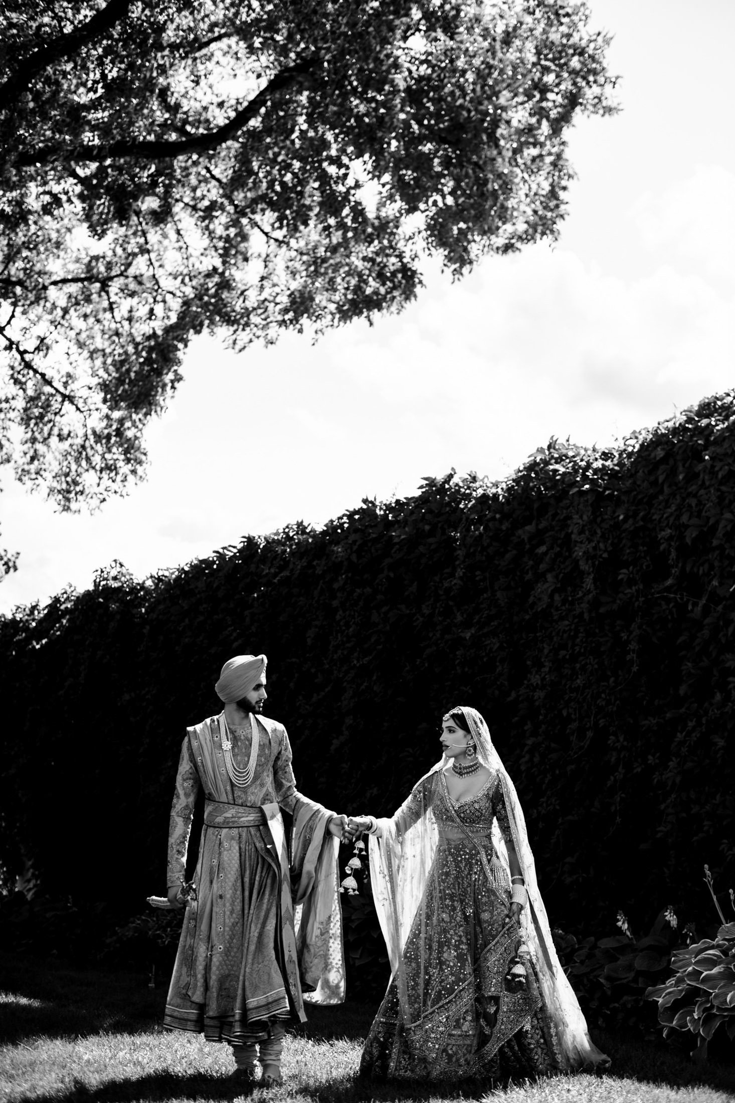 Bride and groom in traditional attire stroll under a grand tree at their Winnipeg wedding.