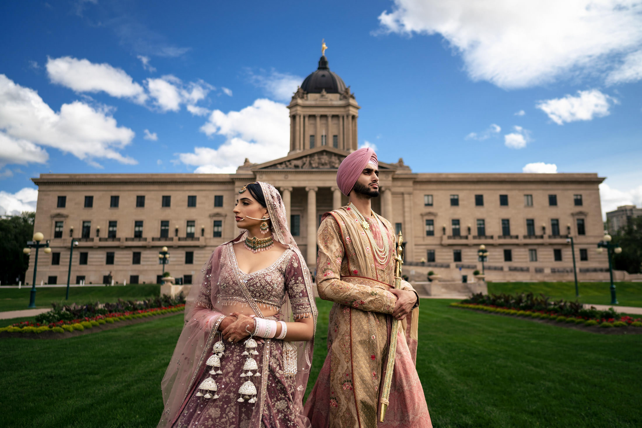 Couple in traditional attire pose at a grand Winnipeg building beneath a cloudy sky.