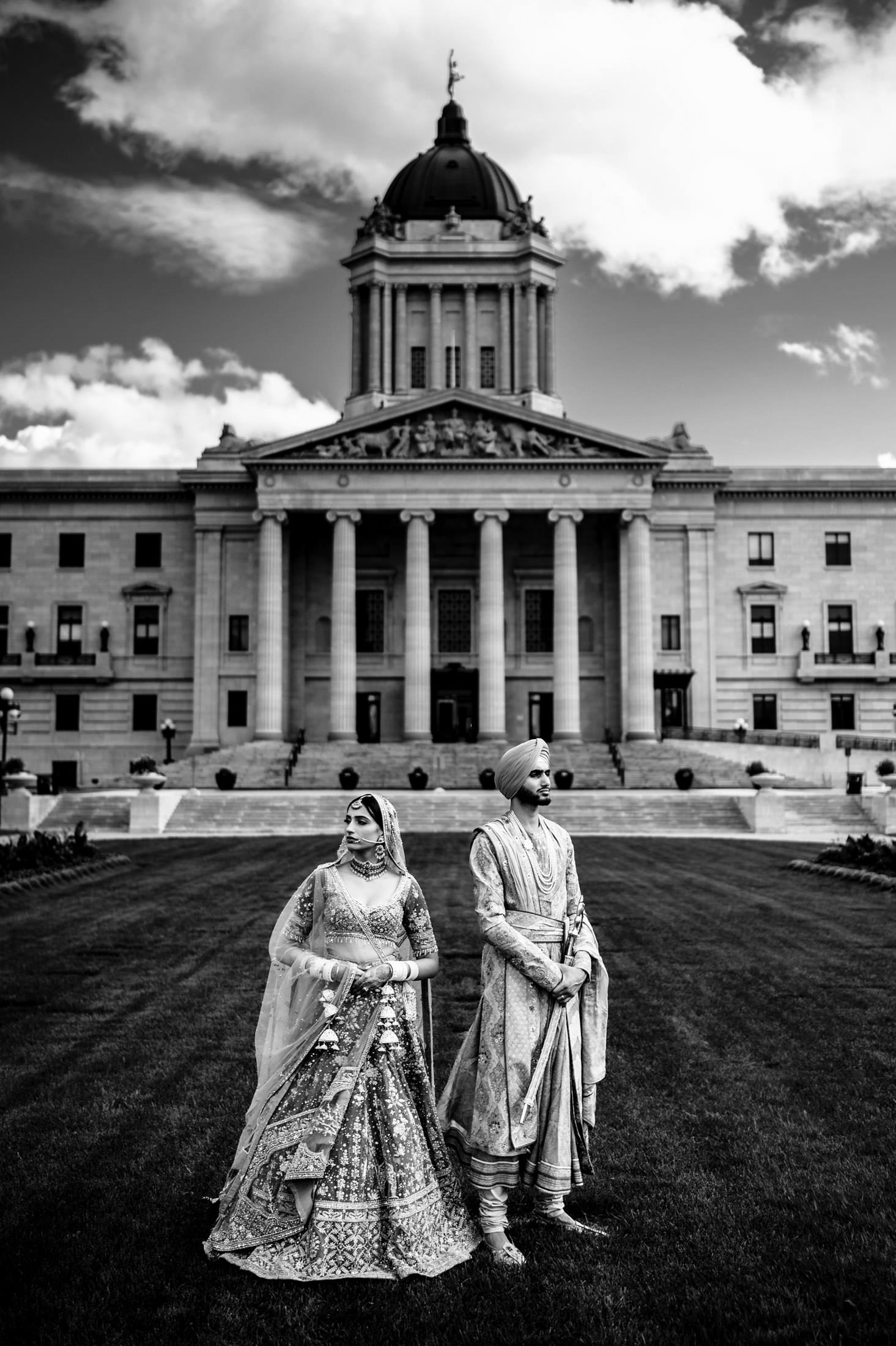 The couple in traditional attire stands before a grand Winnipeg wedding venue.