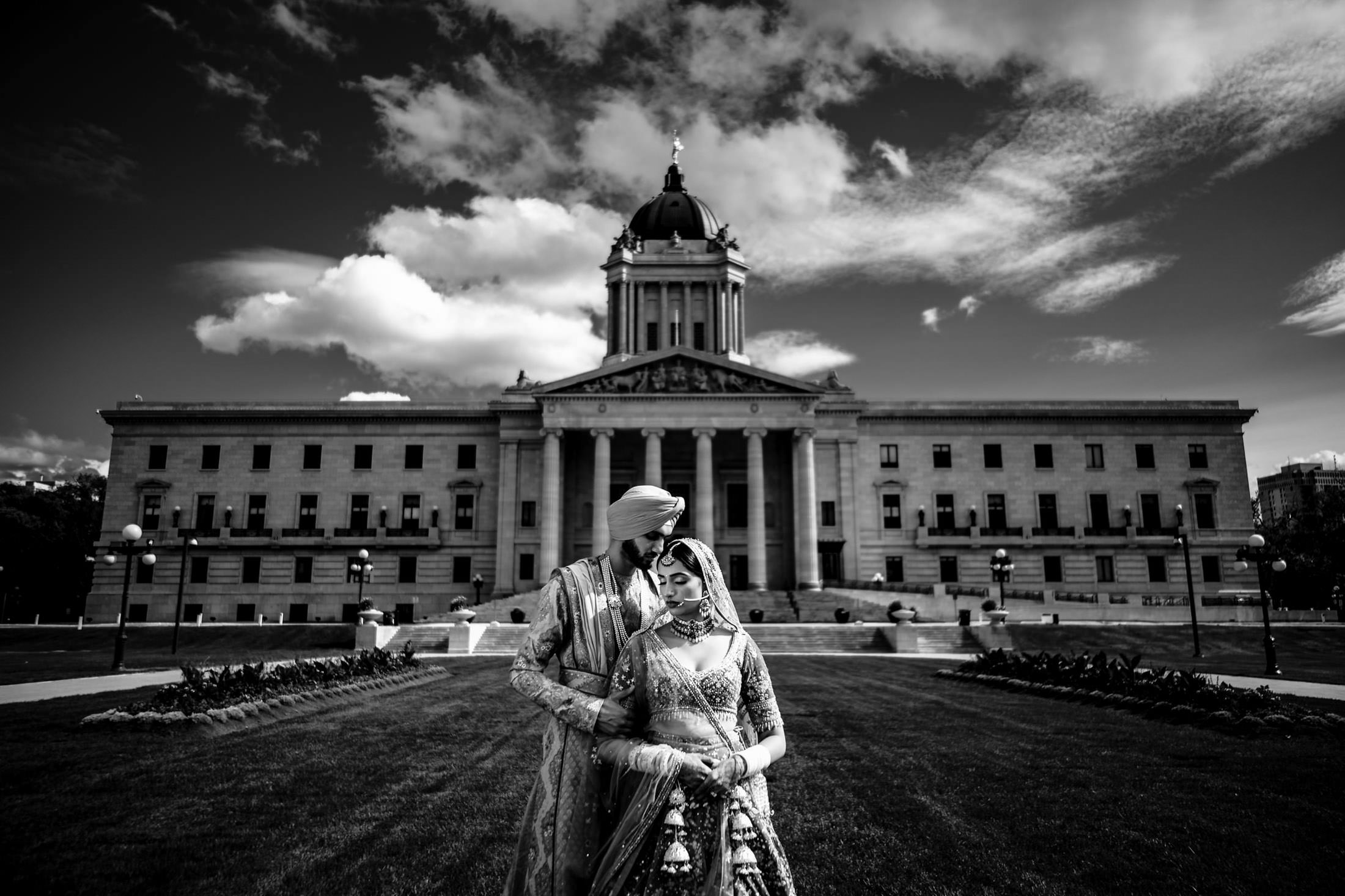 Bride and groom in traditional attire pose at a grand Winnipeg wedding site under cloudy skies.