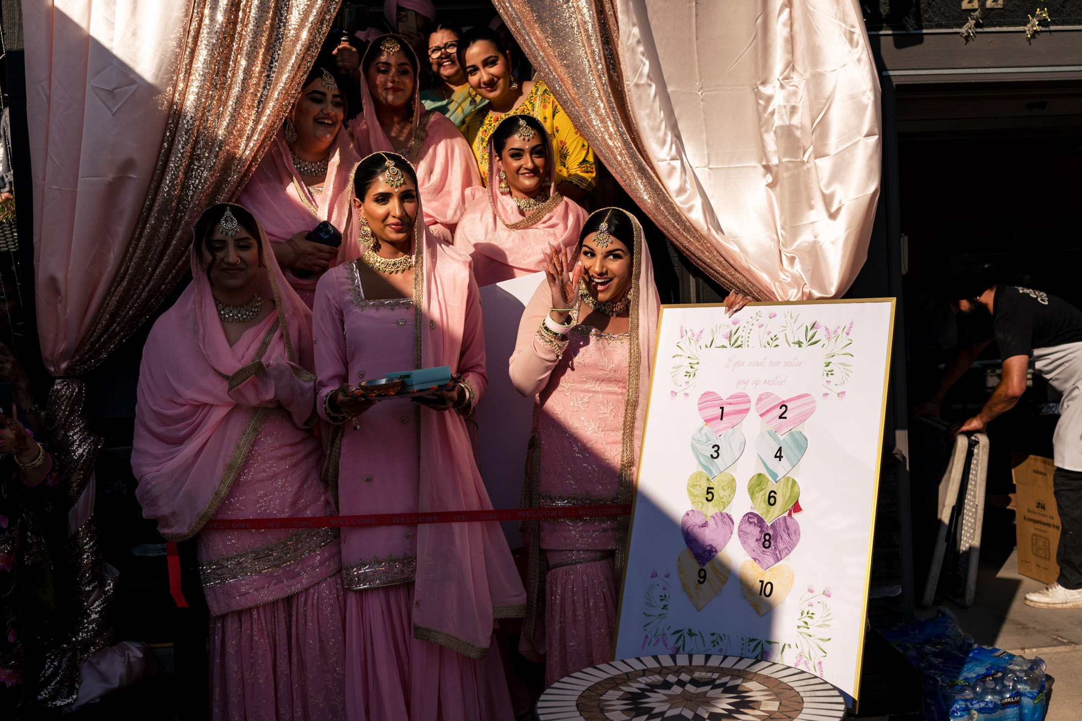 Women in pink traditional attire pose by a heart-decorated board at a Winnipeg wedding.