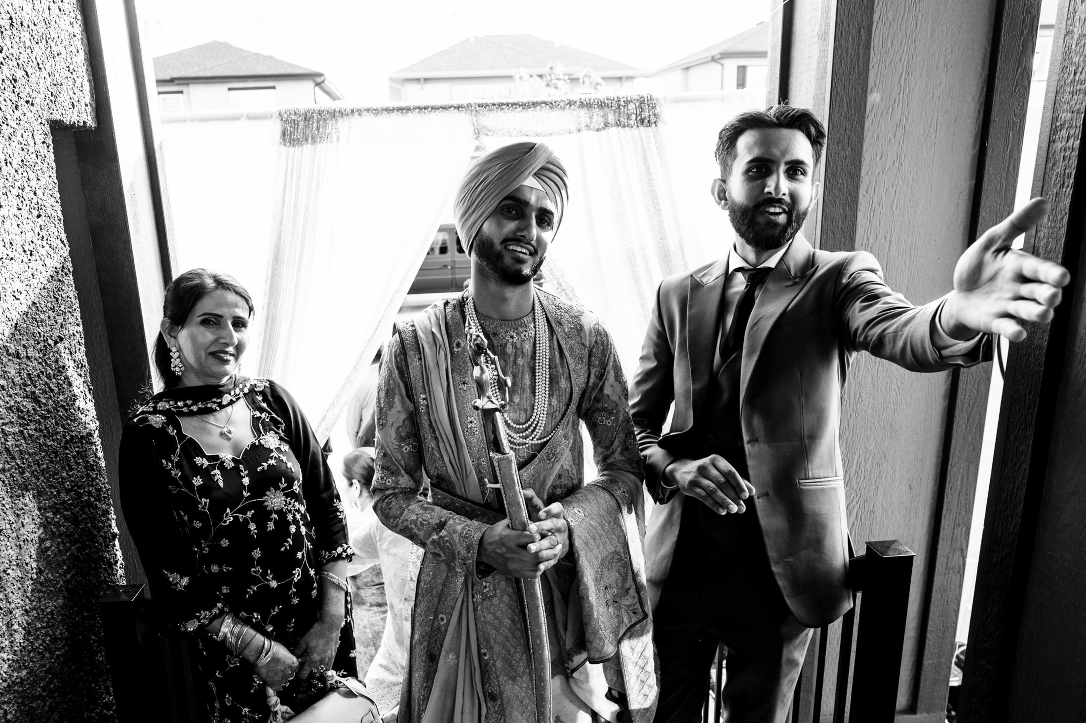 Three people in formal attire, one in traditional clothing, smile at a Winnipeg wedding outdoors.