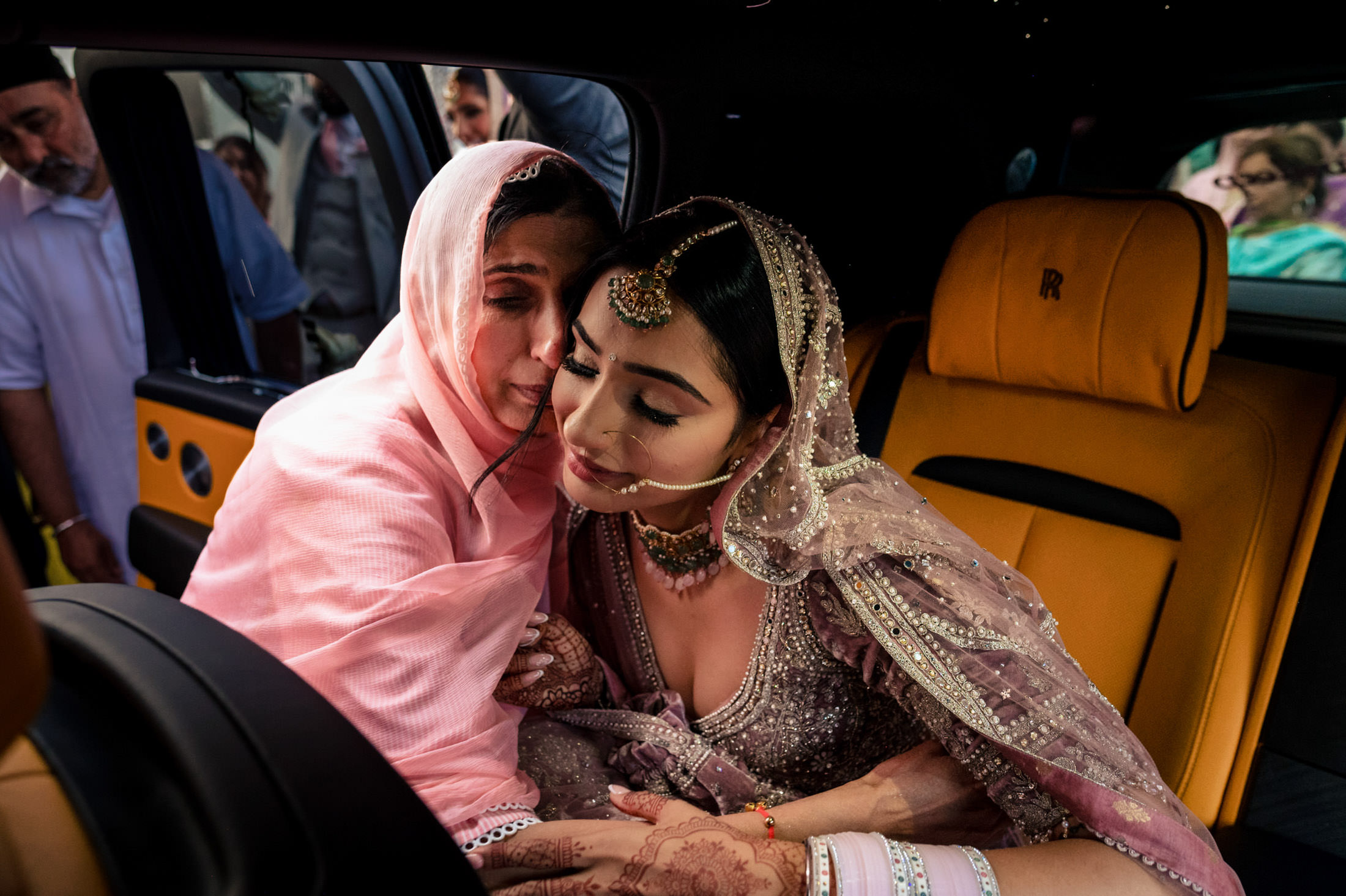 Bride and woman share a heartfelt moment in a car at the Winnipeg wedding, surrounded by people.