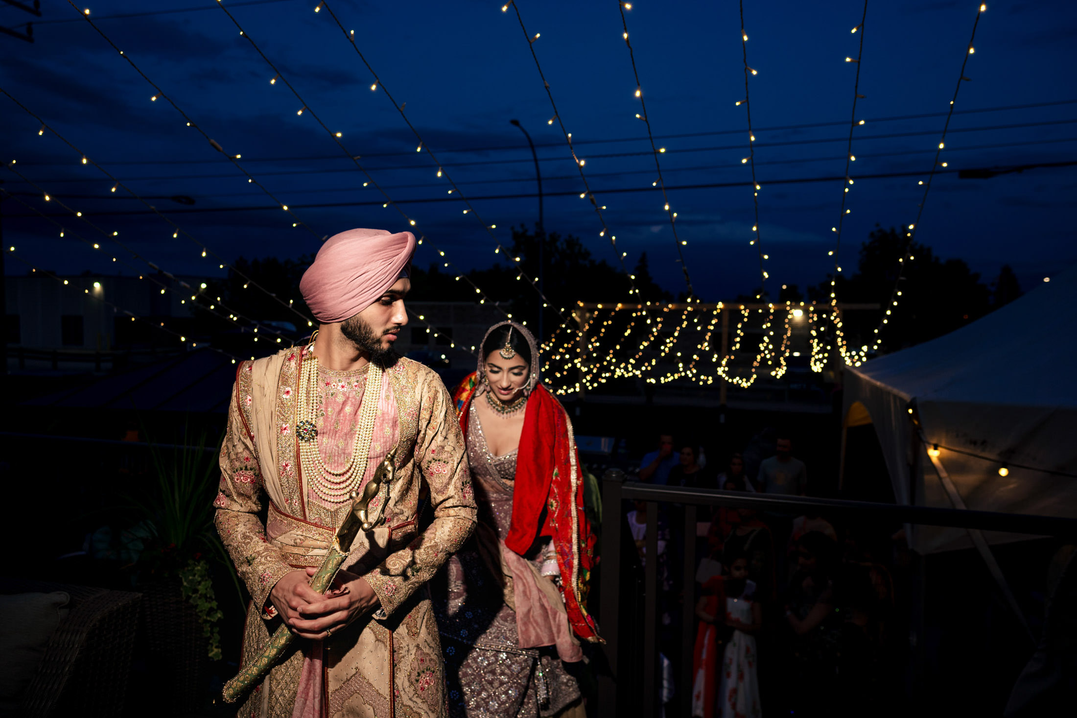 Couple in traditional attire walking under Winnipeg wedding string lights at night.