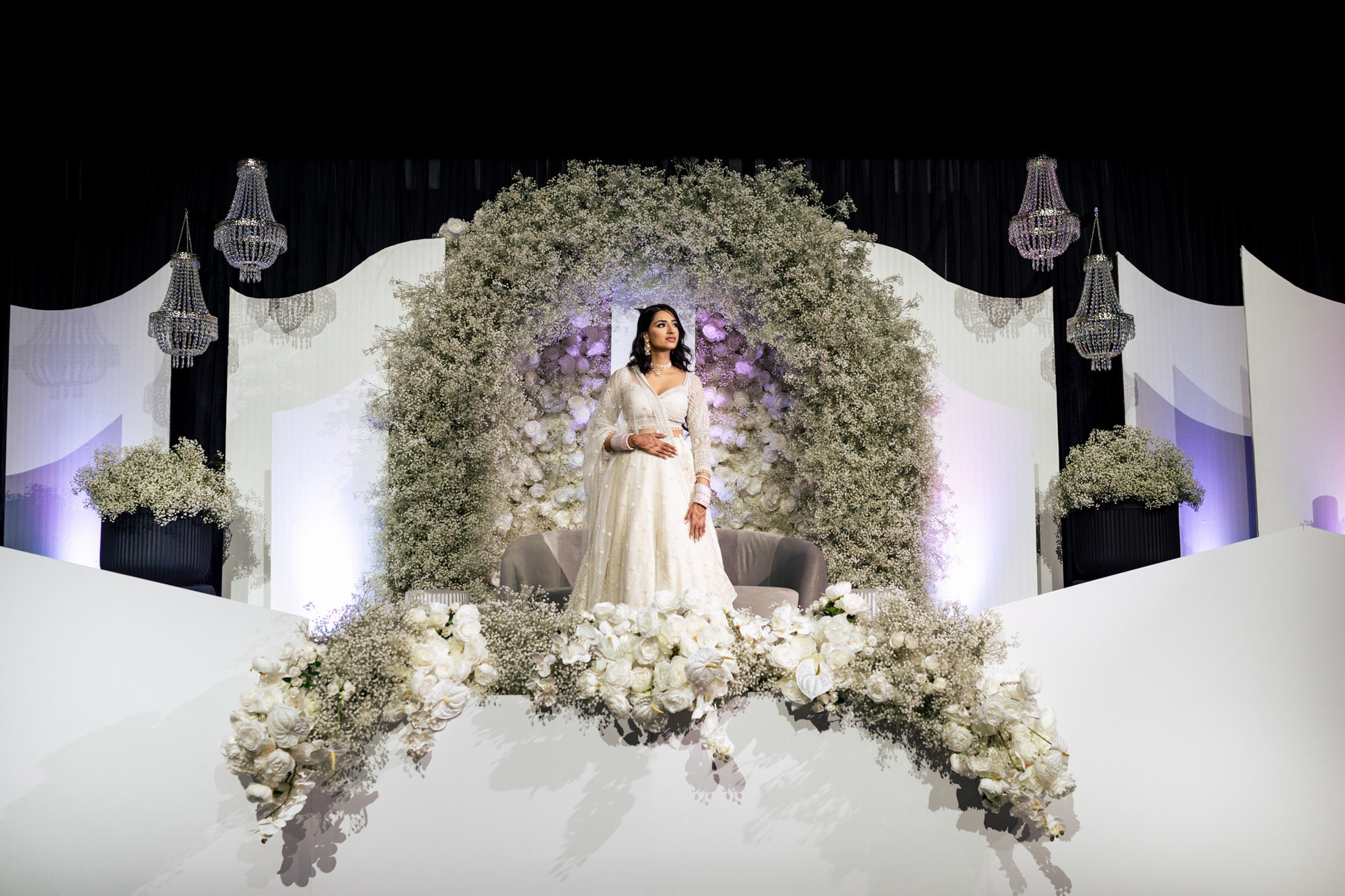 Bride in white gown seated in floral arch with chandeliers at a Winnipeg wedding.