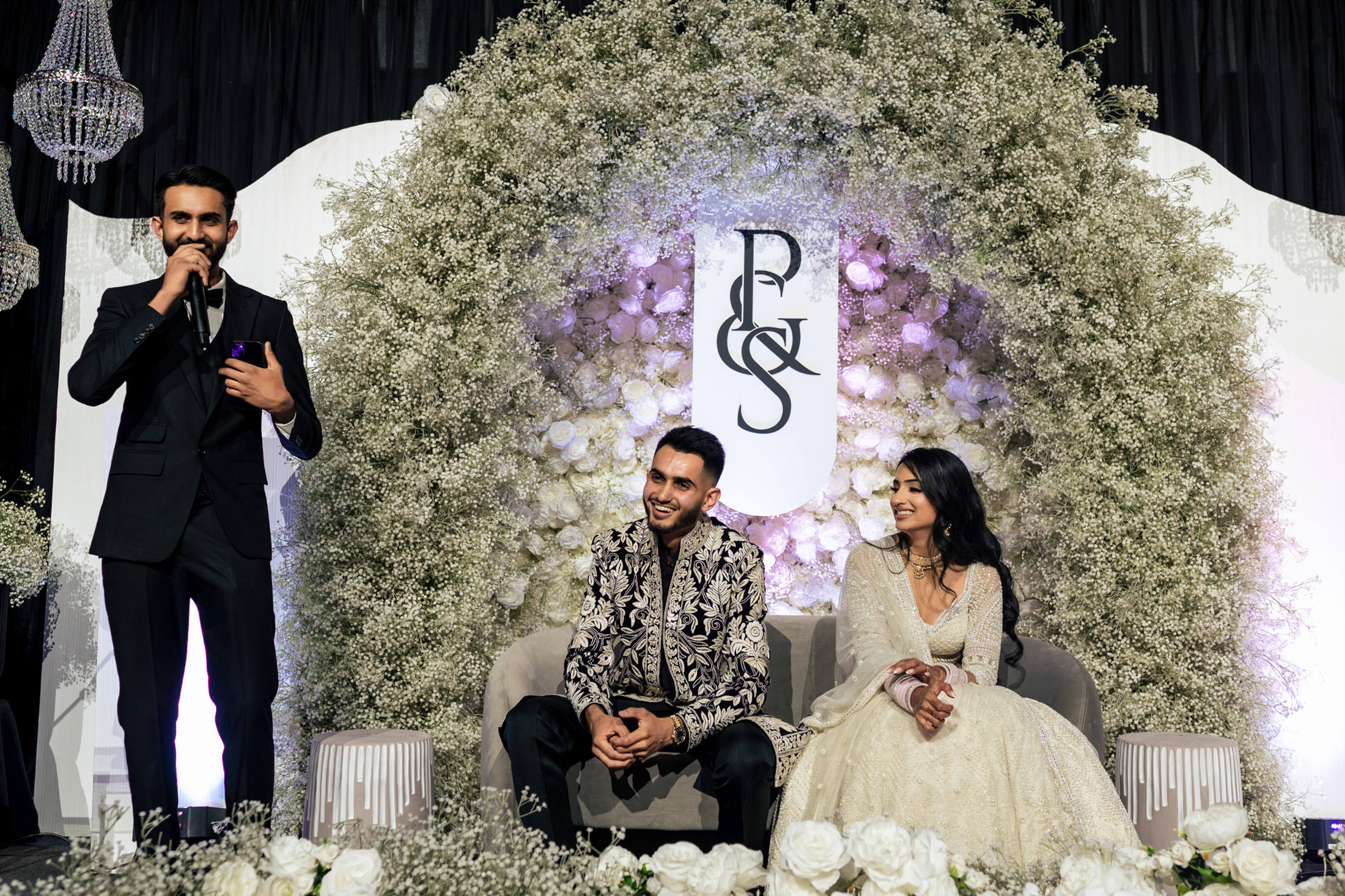 Man speaking at Winnipeg wedding, couple seated, floral backdrop with initials "P & S.