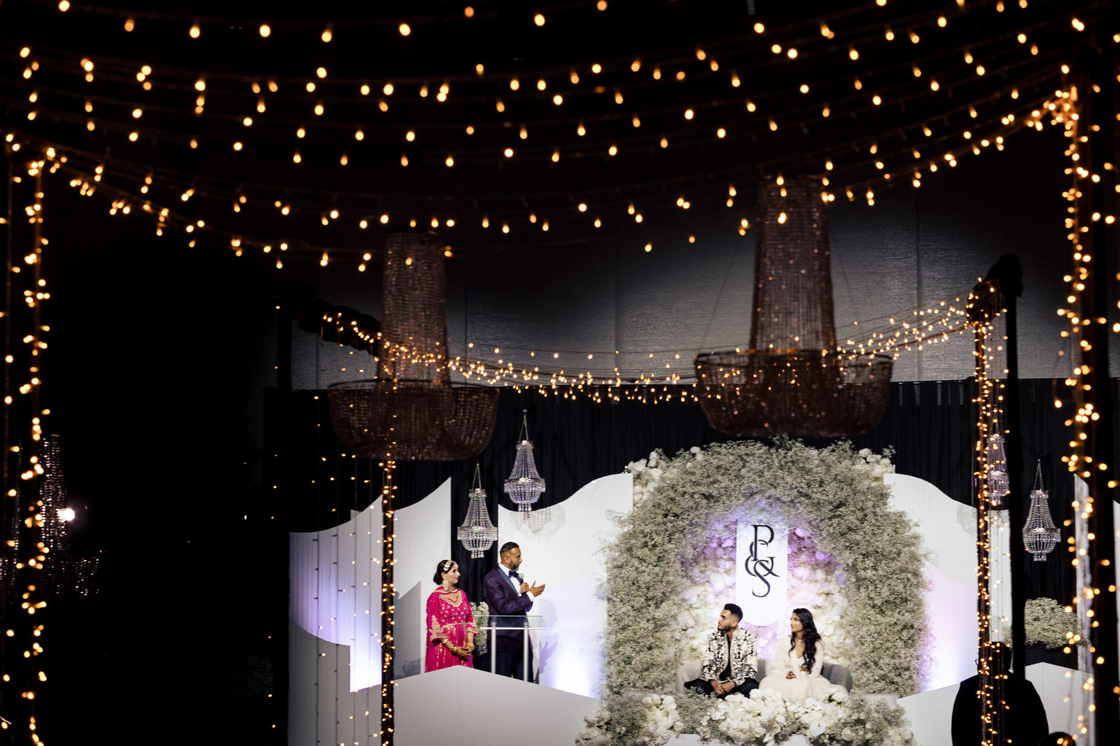 A couple sits on stage under fairy lights during a Winnipeg wedding, as two speak at a podium.