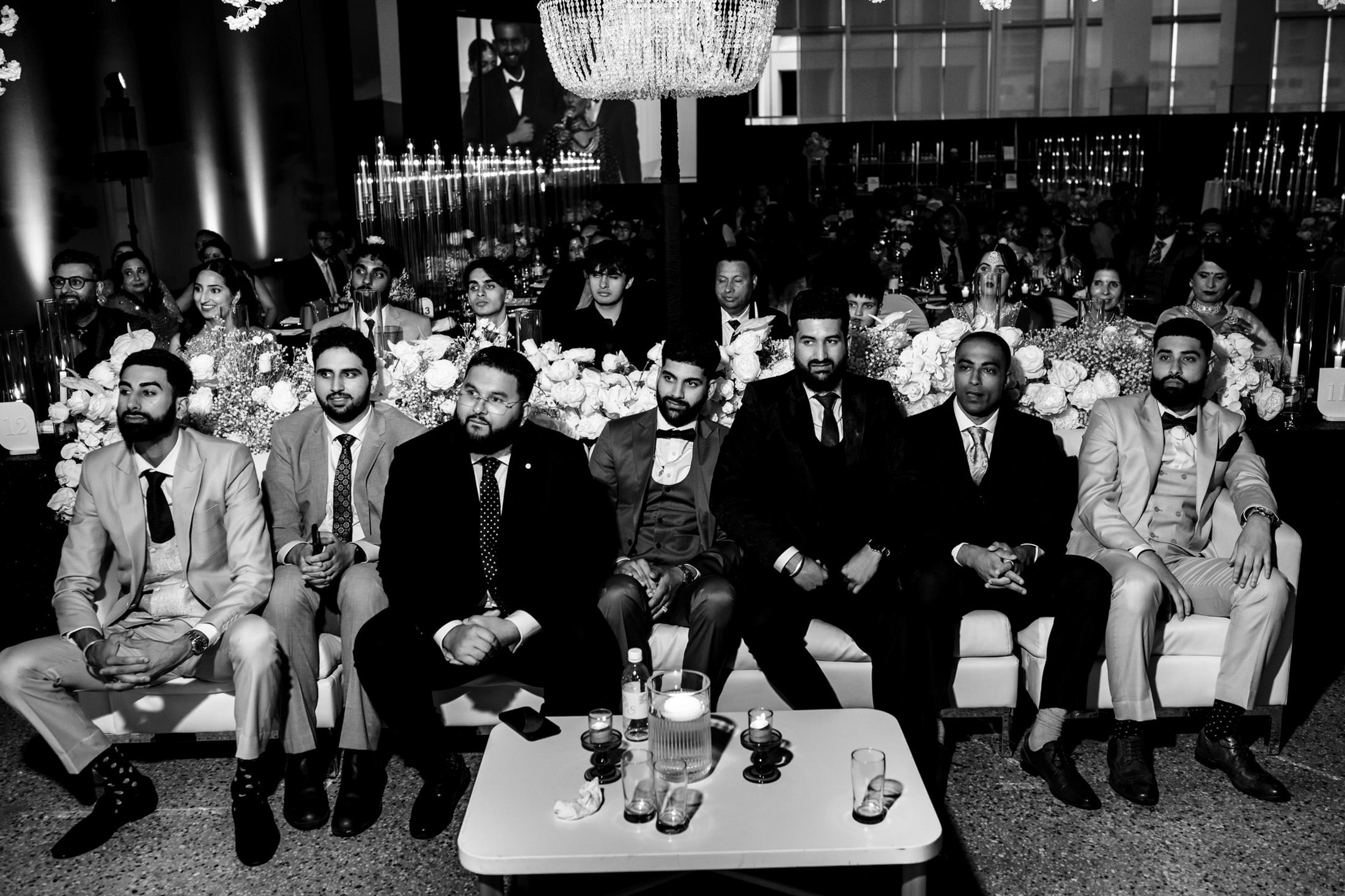 Men in suits seated together at a Winnipeg wedding, with a crowd behind them.