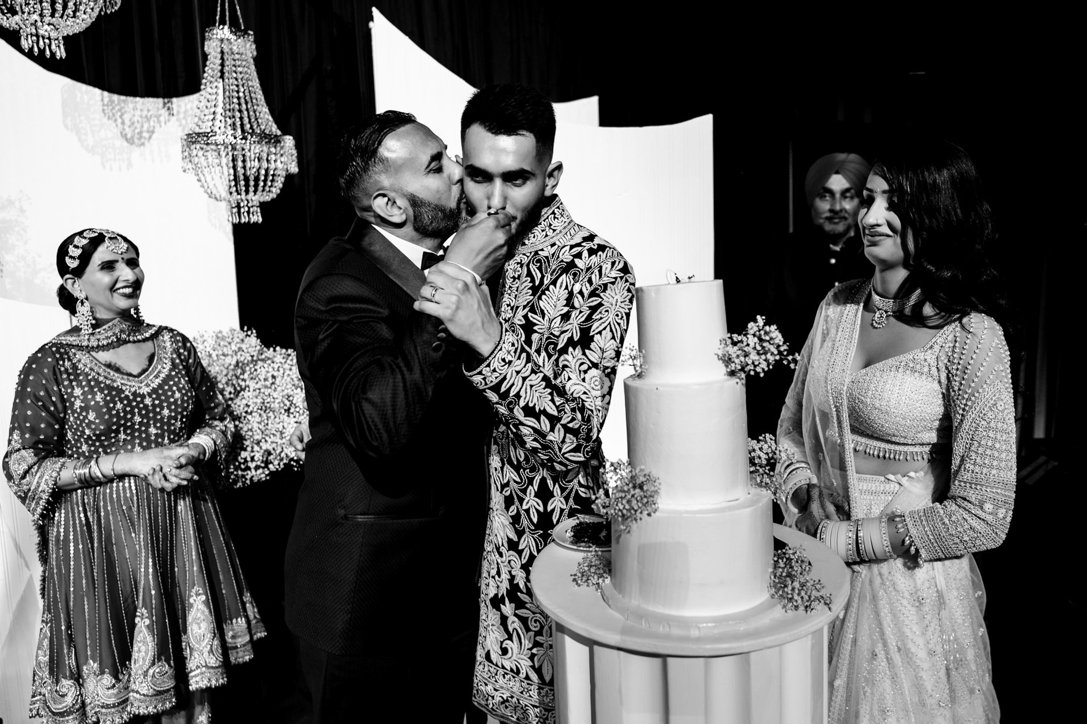 A couple shares a kiss by a cake at their Winnipeg wedding, surrounded by joyful guests in traditional attire.