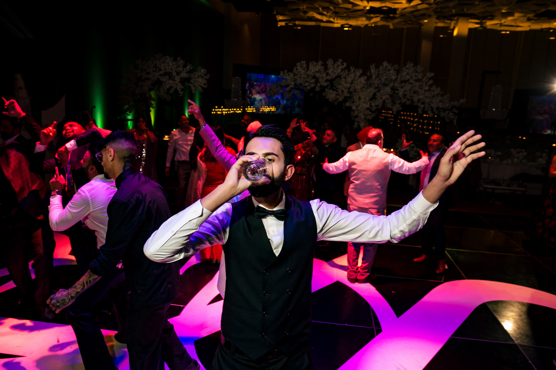 Person in formal wear sipping at a lively Winnipeg wedding, dancers in the background.