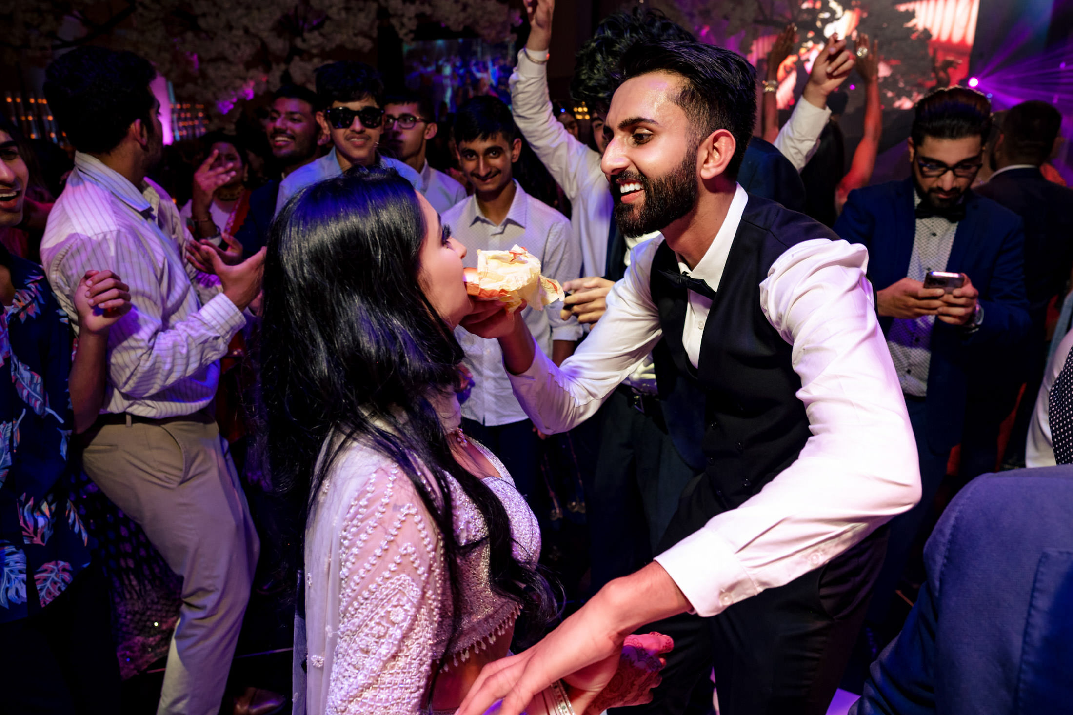 At a lively Winnipeg wedding, a man feeds cake to a woman amid dancing guests.