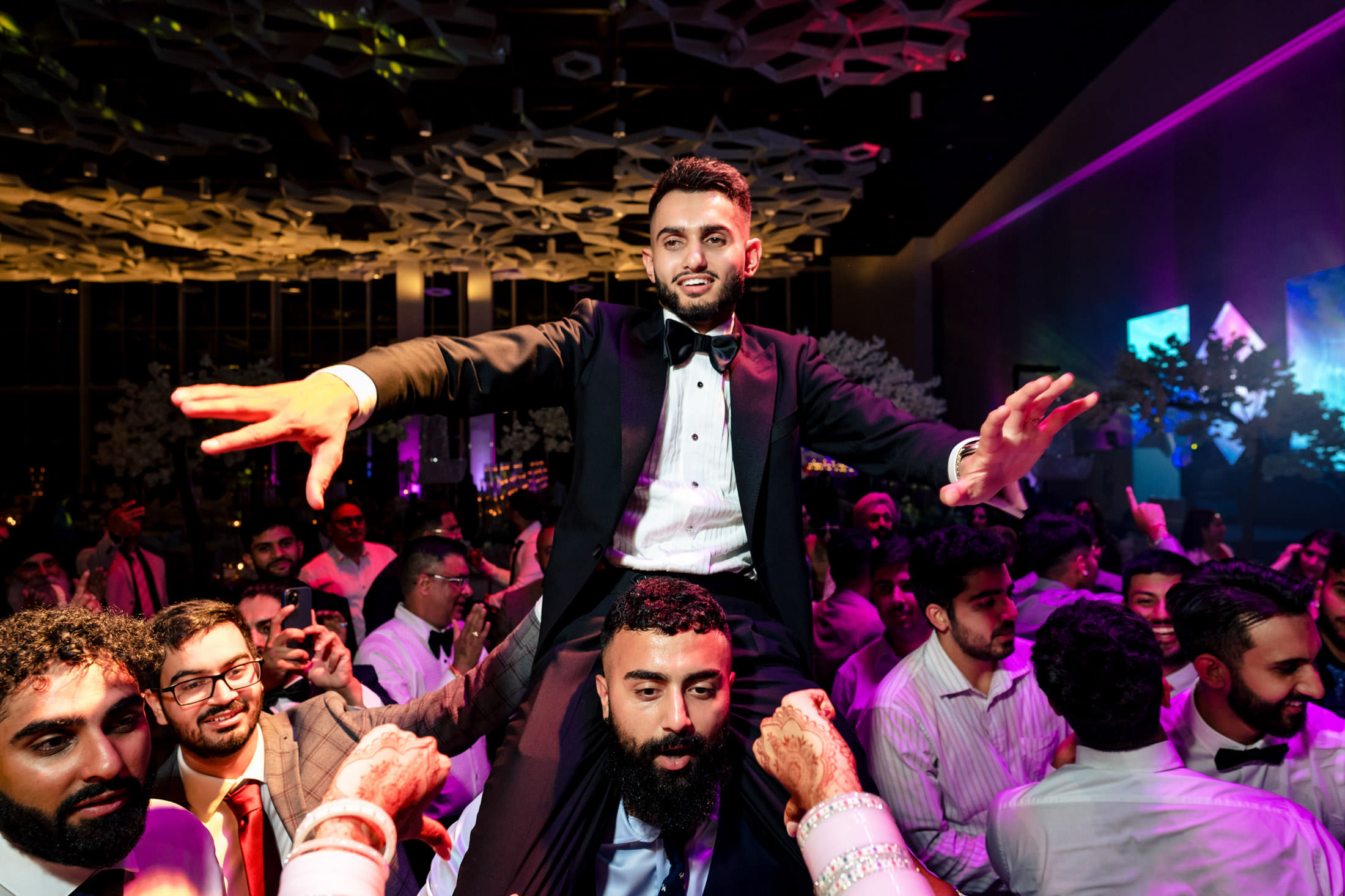 Man in tuxedo lifted on shoulders at lively Winnipeg wedding with colorful lights.
