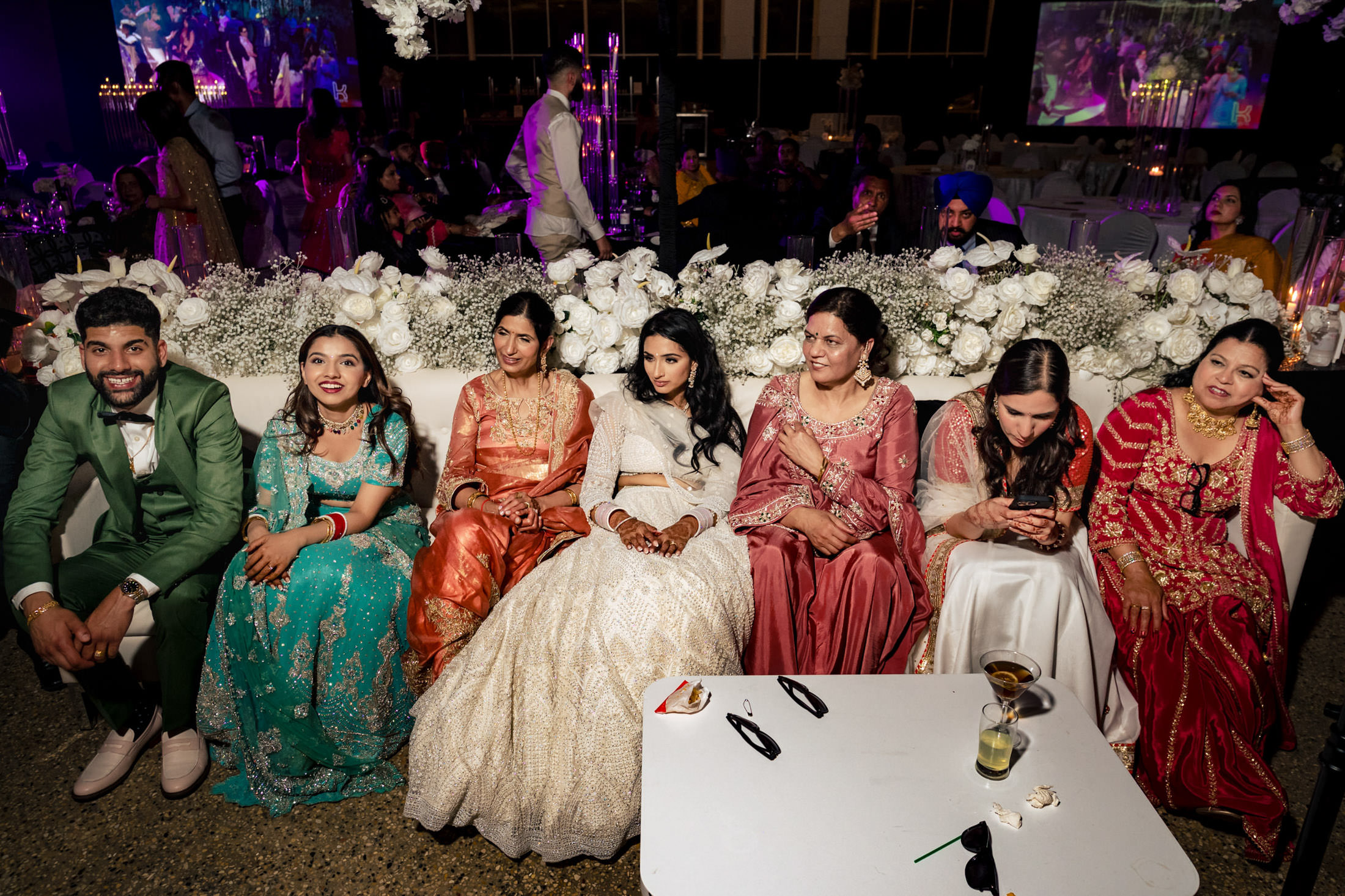 Group seated in colorful attire at a Winnipeg wedding, with florals and screens behind.