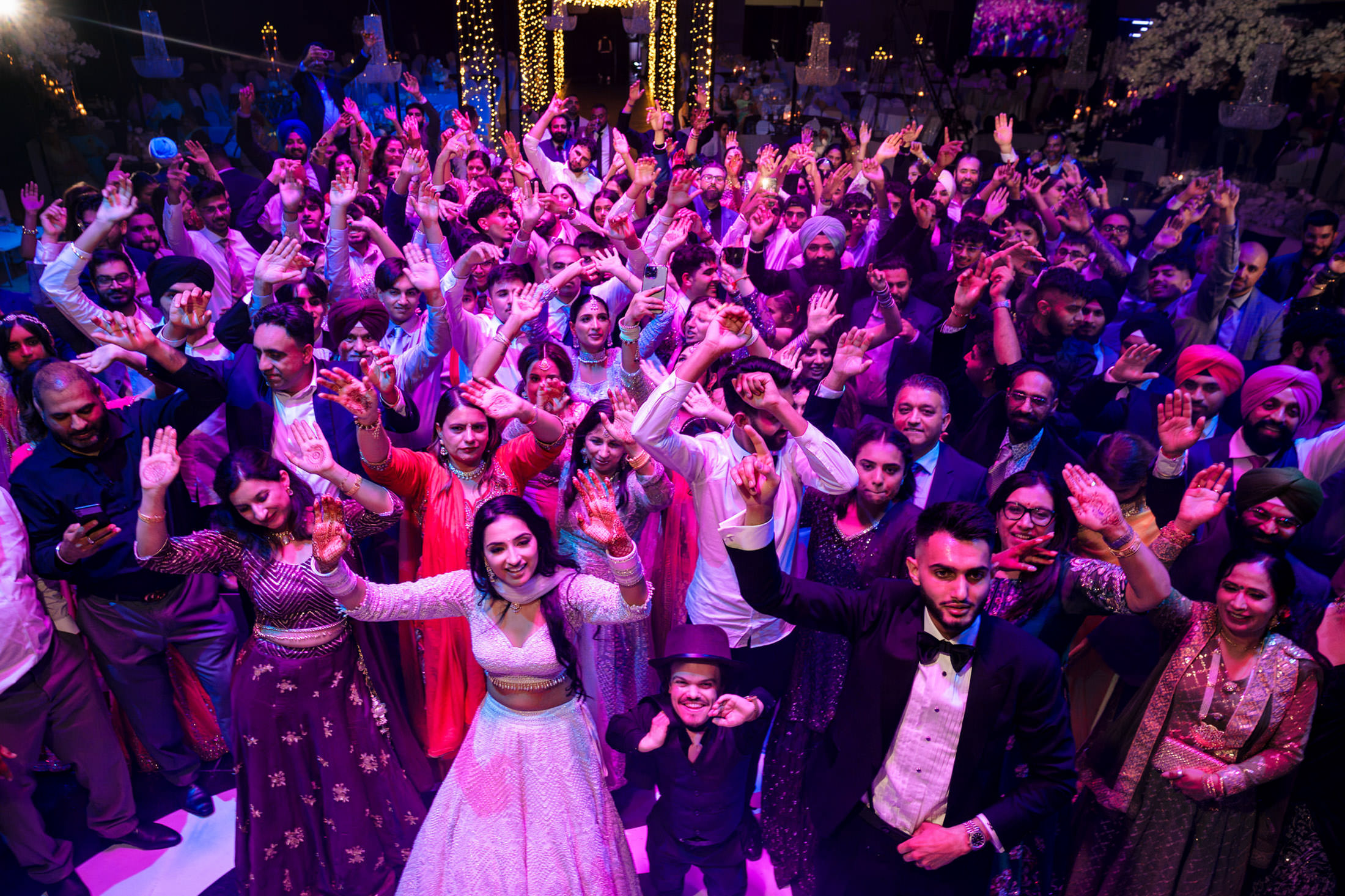 A large group joyfully dancing at a vibrant Winnipeg wedding under colorful lights.