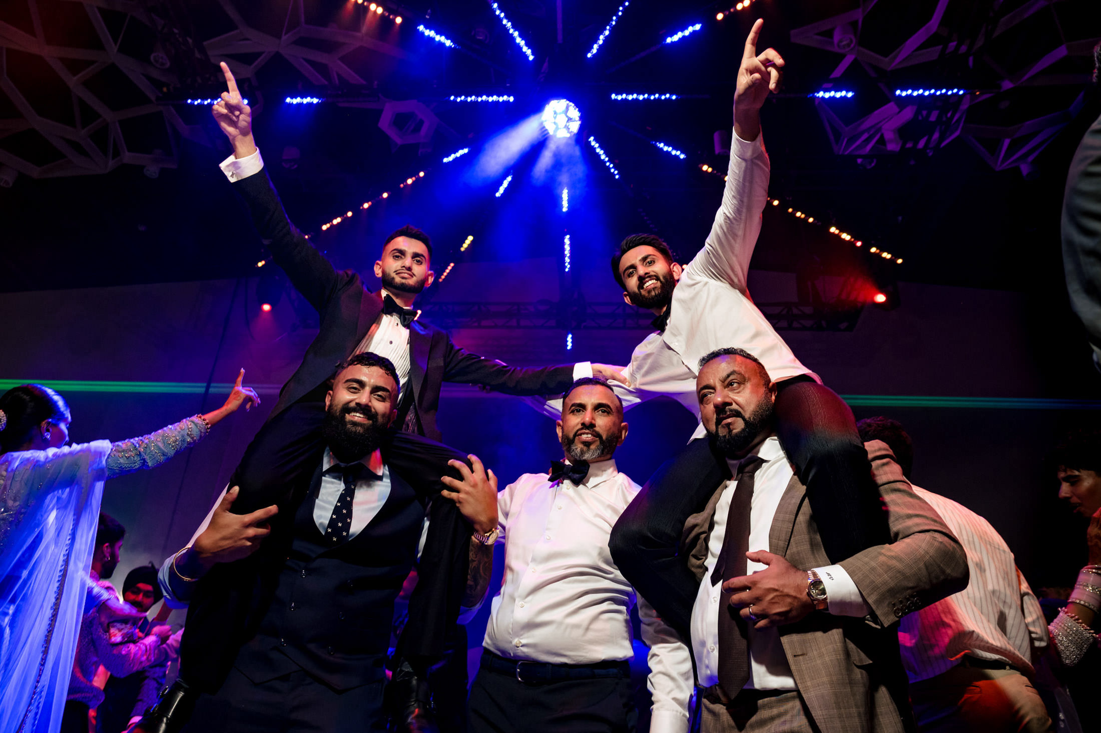 Men in suits celebrate at a Winnipeg wedding, dancing with two on shoulders under colorful lights.