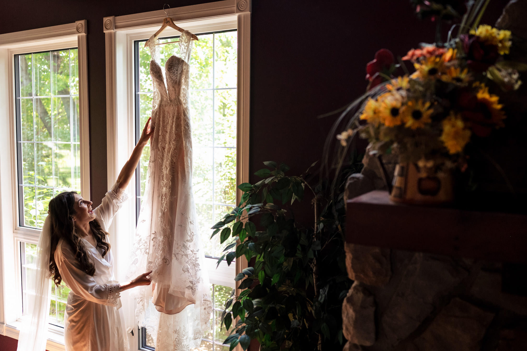 Woman reaching for a hanging wedding dress by a window at Ashgrove Acres, flowers nearby.