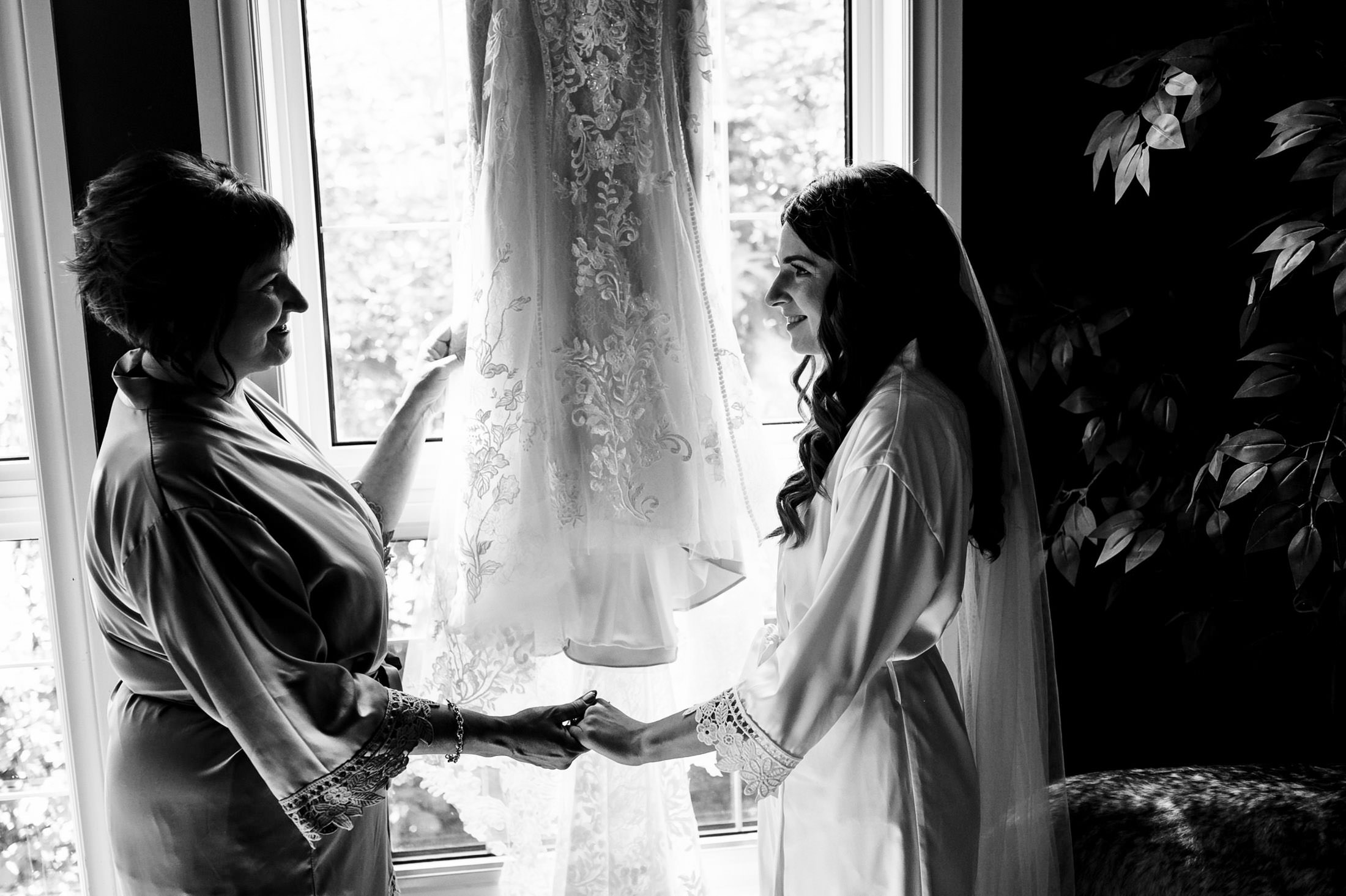 Two women holding hands near a window at Ashgrove Acres, a lace wedding dress between them.