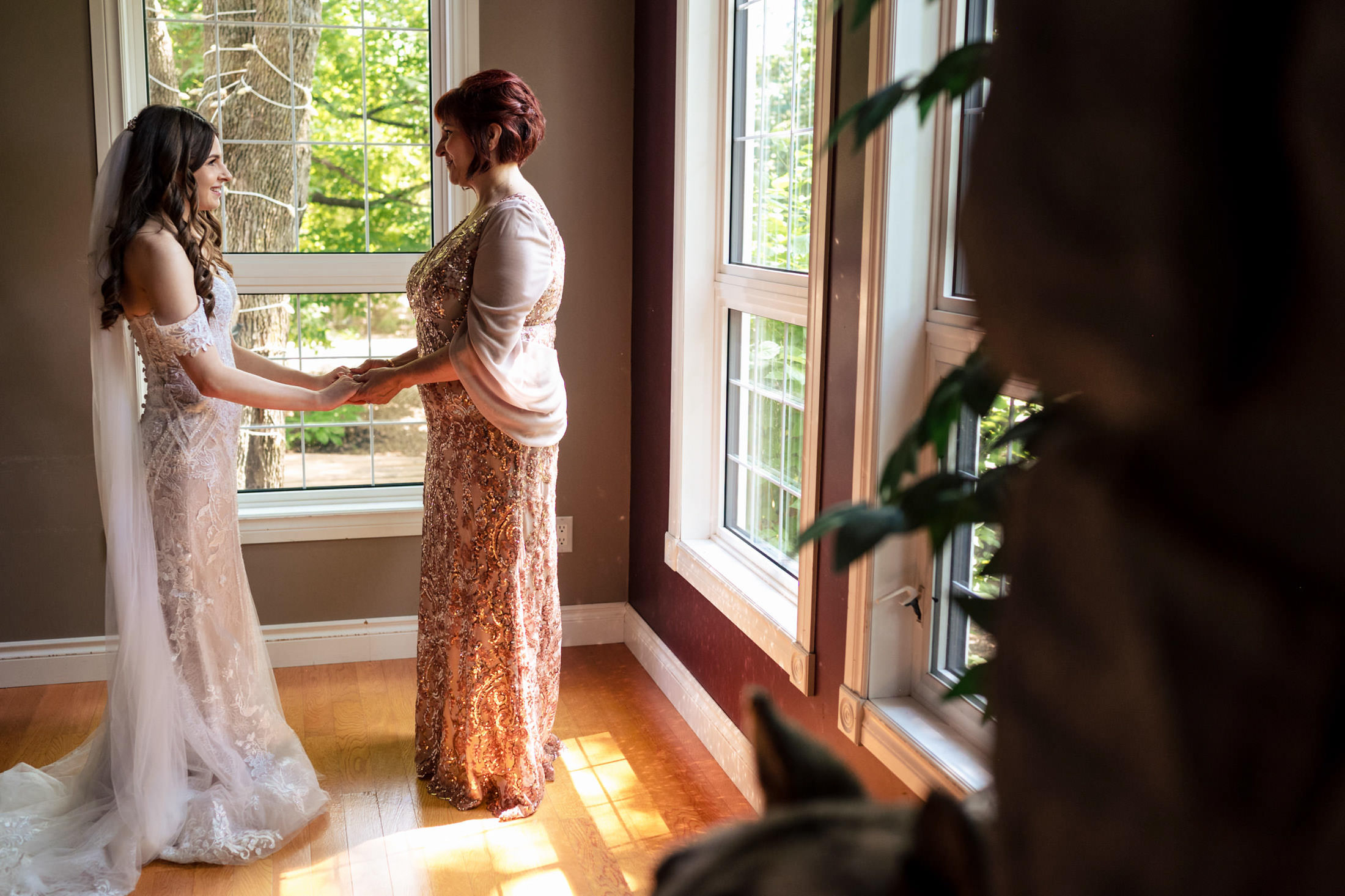 Bride and woman holding hands by a sunlit window at Ashgrove Acres with trees outside.