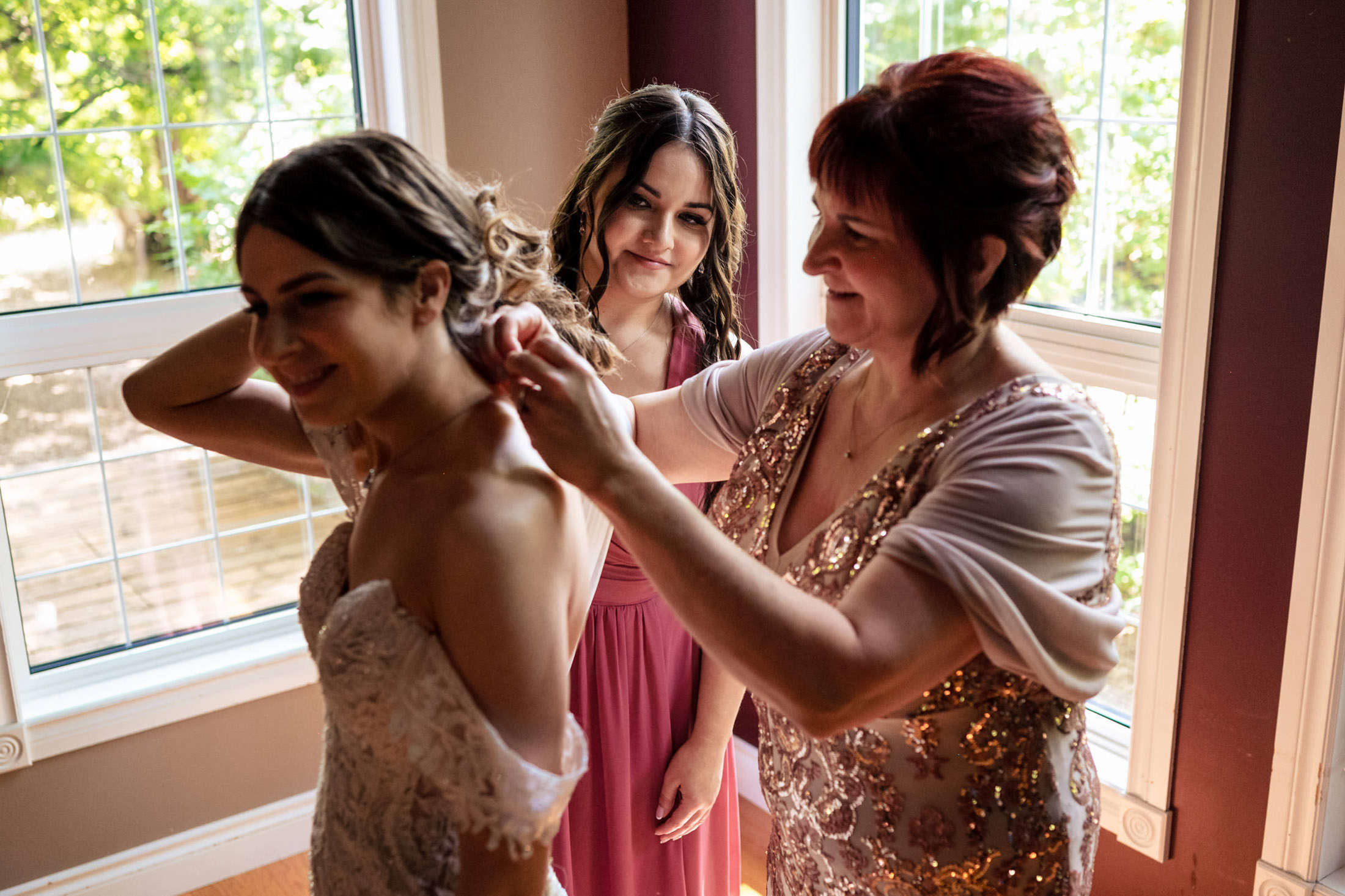 Bride getting ready by the window at Ashgrove Acres, aided by two women with her dress.