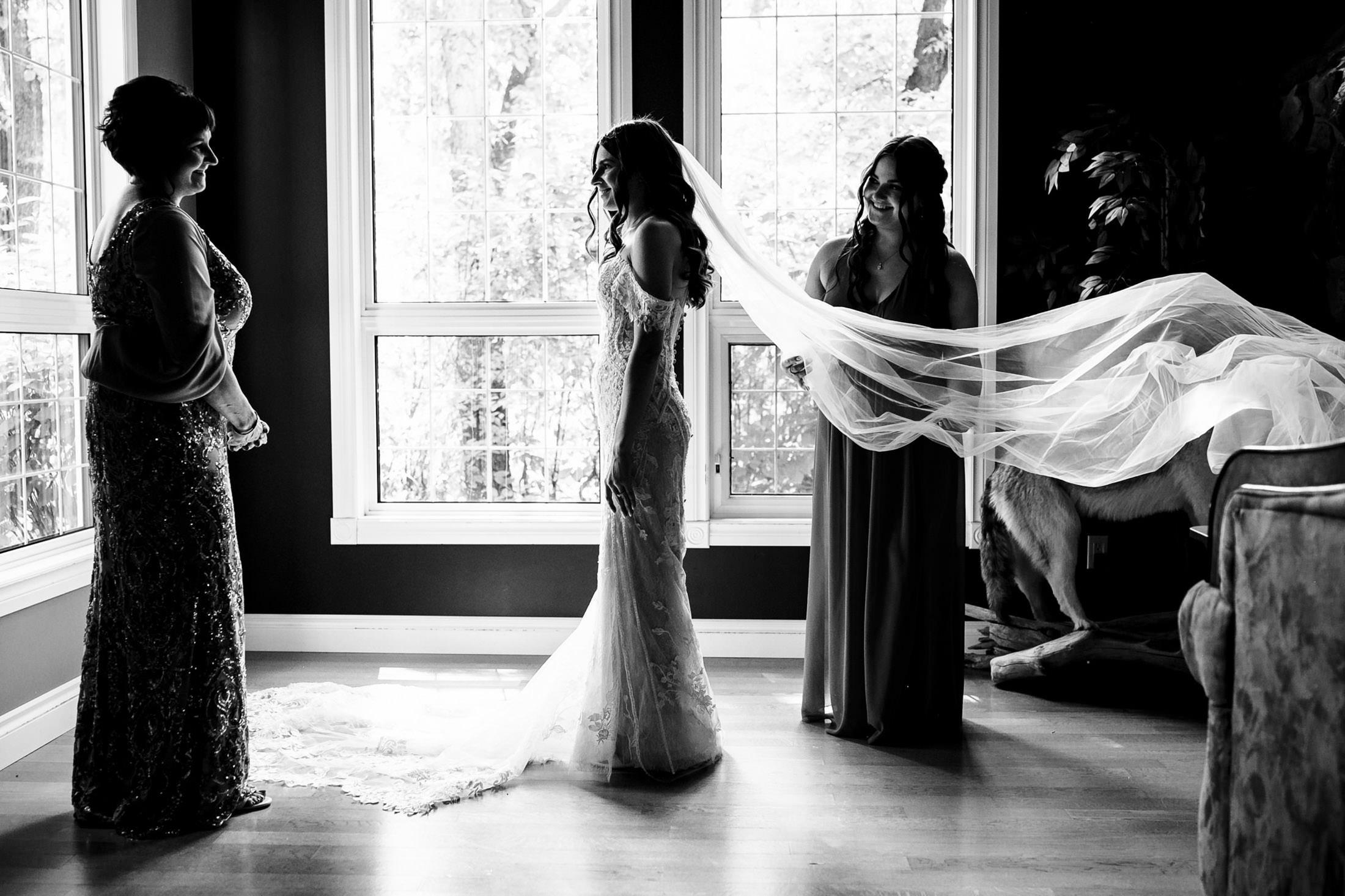 Bride in gown with veil adjusted by bridesmaid at Ashgrove Acres, woman looks on in sunlit room.