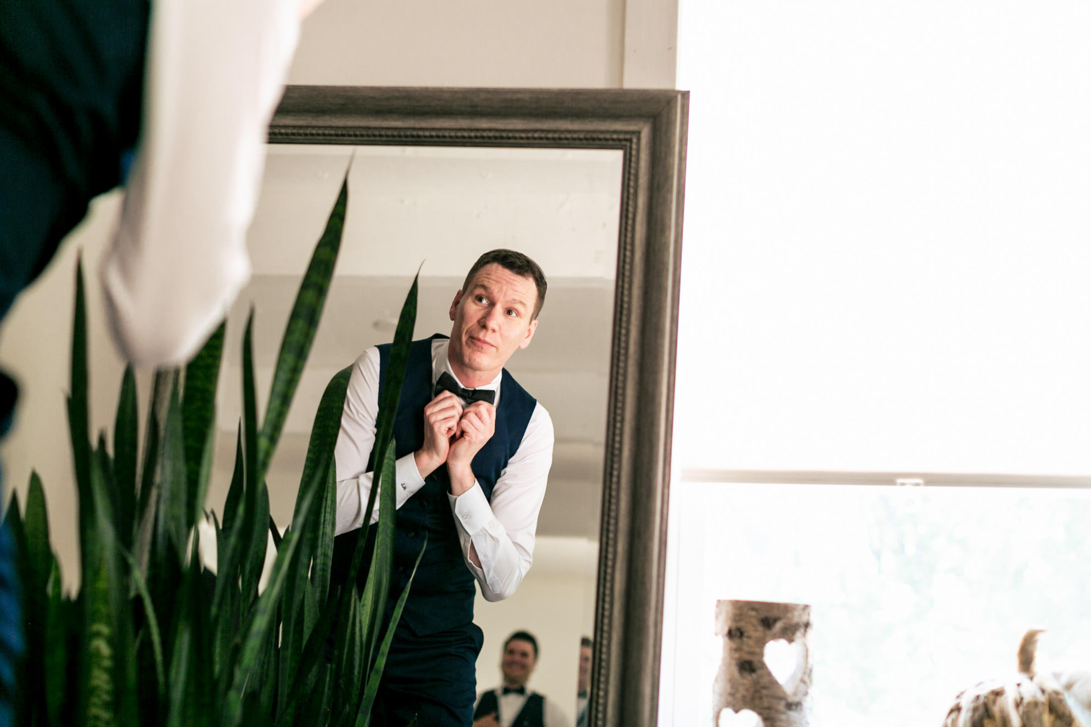 Man adjusting bow tie for Ashgrove Acres wedding, wearing a vest, with plants in the foreground.