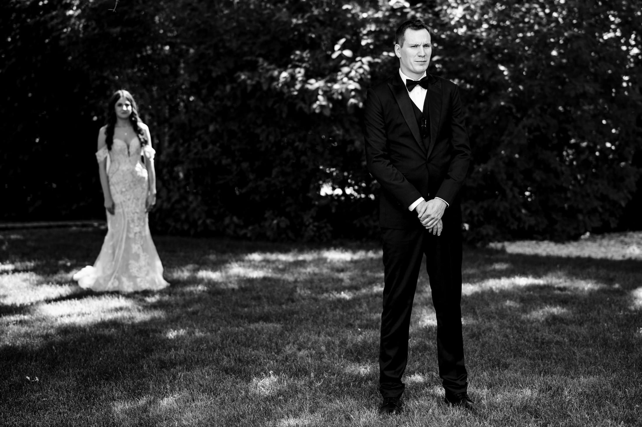 Bride approaches groom on grass at Ashgrove Acres. Sunlit garden, black and white photo.