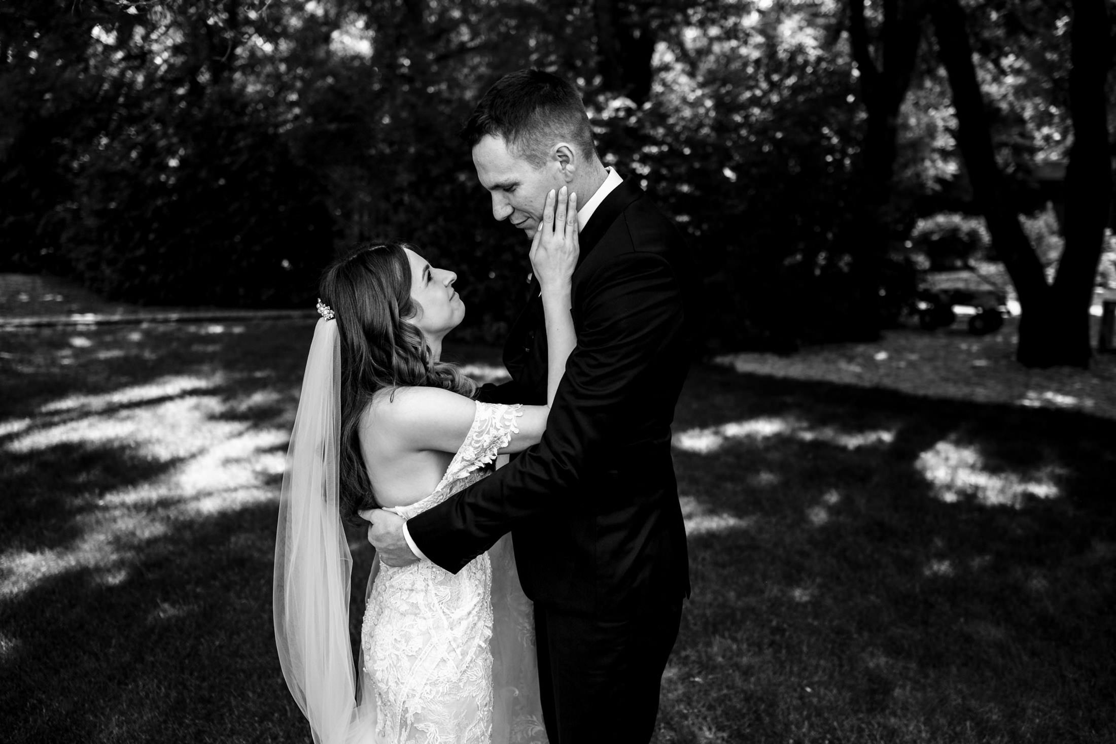 Bride and groom embrace at Ashgrove Acres, gazing in a sunlit, wooded area.
