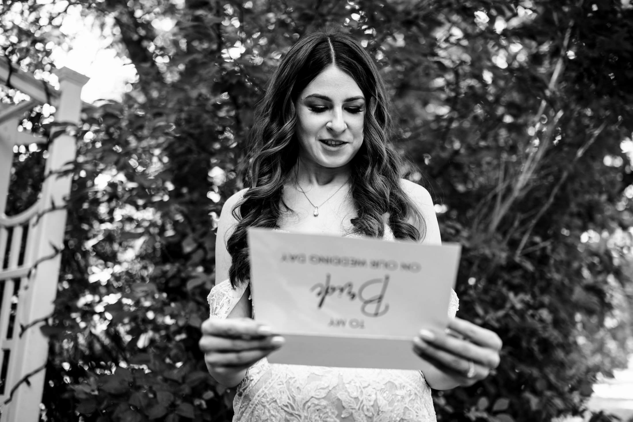 Bride smiles at card outdoors, surrounded by trees and garden trellis at Ashgrove Acres.
