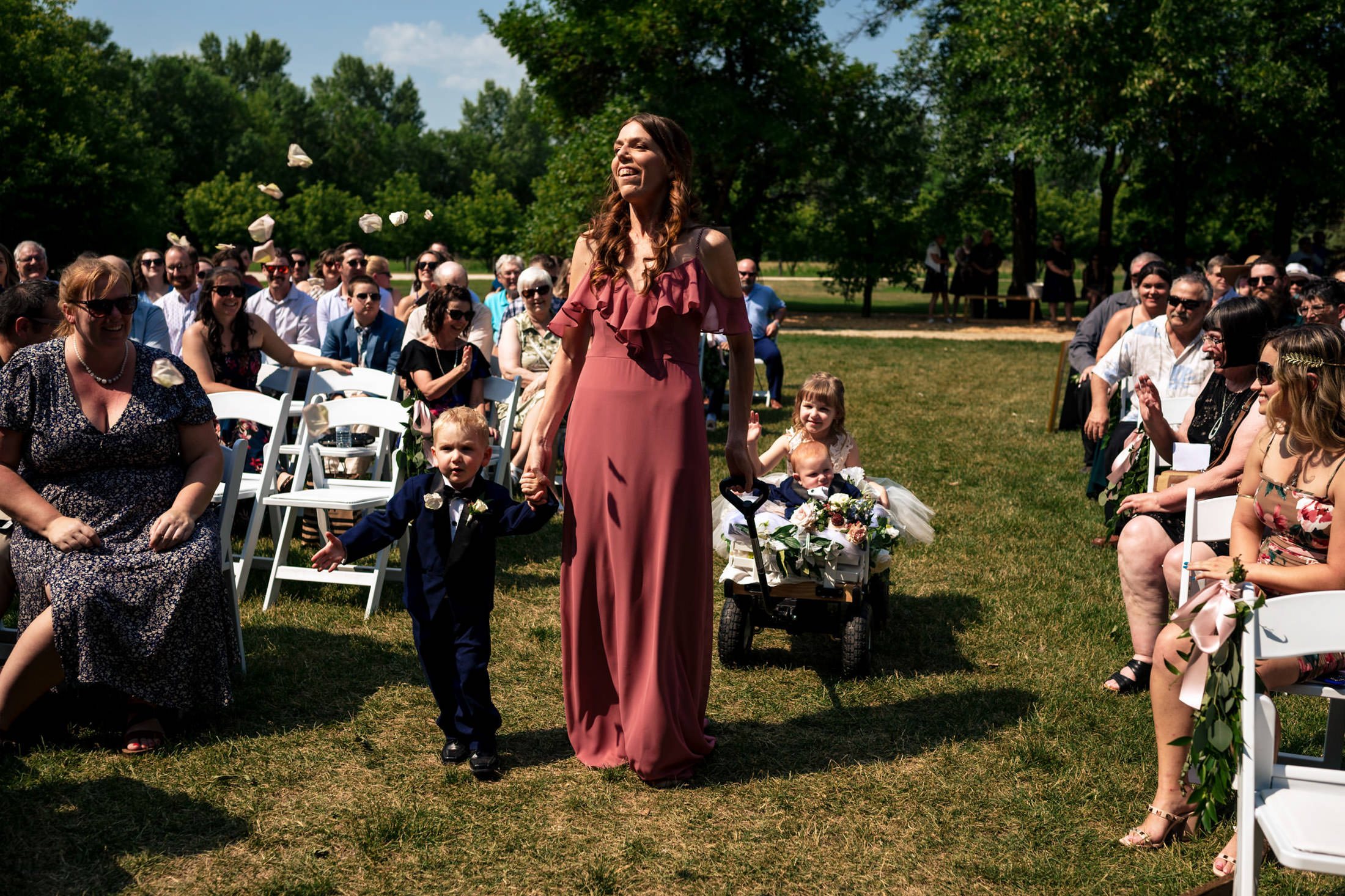 At Ashgrove Acres Wedding, a woman and boy stroll down the aisle, pulling a wagon with two kids.