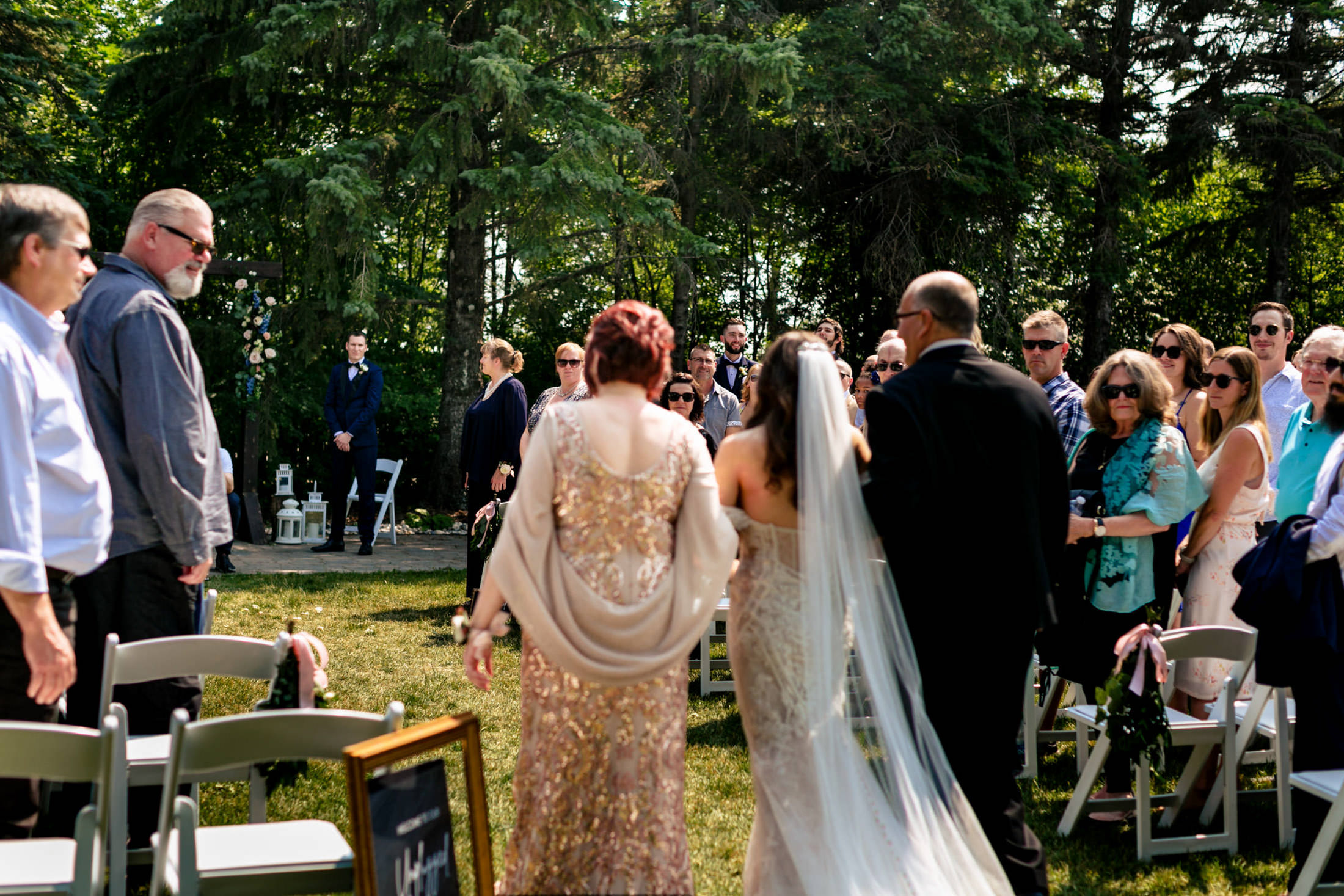 At Ashgrove Acres, the bride walks down the aisle with parents amid seated guests and lush trees.